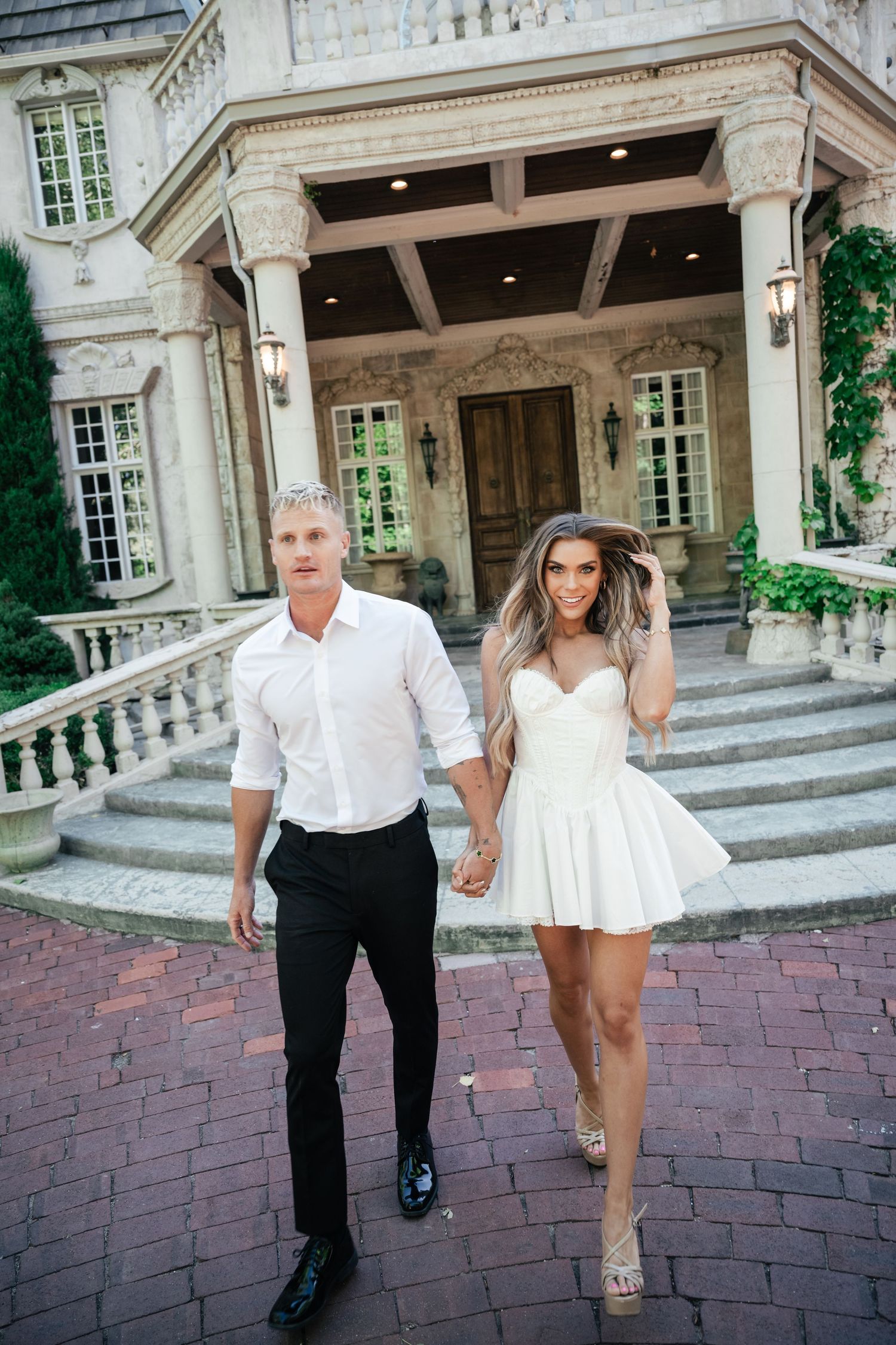 A couple in elegant white attire walking down brick steps in front of a luxurious mansion entrance.