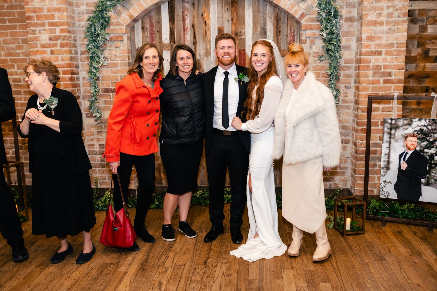 A group stands together at a wedding reception venue with exposed brick walls and greenery decor.