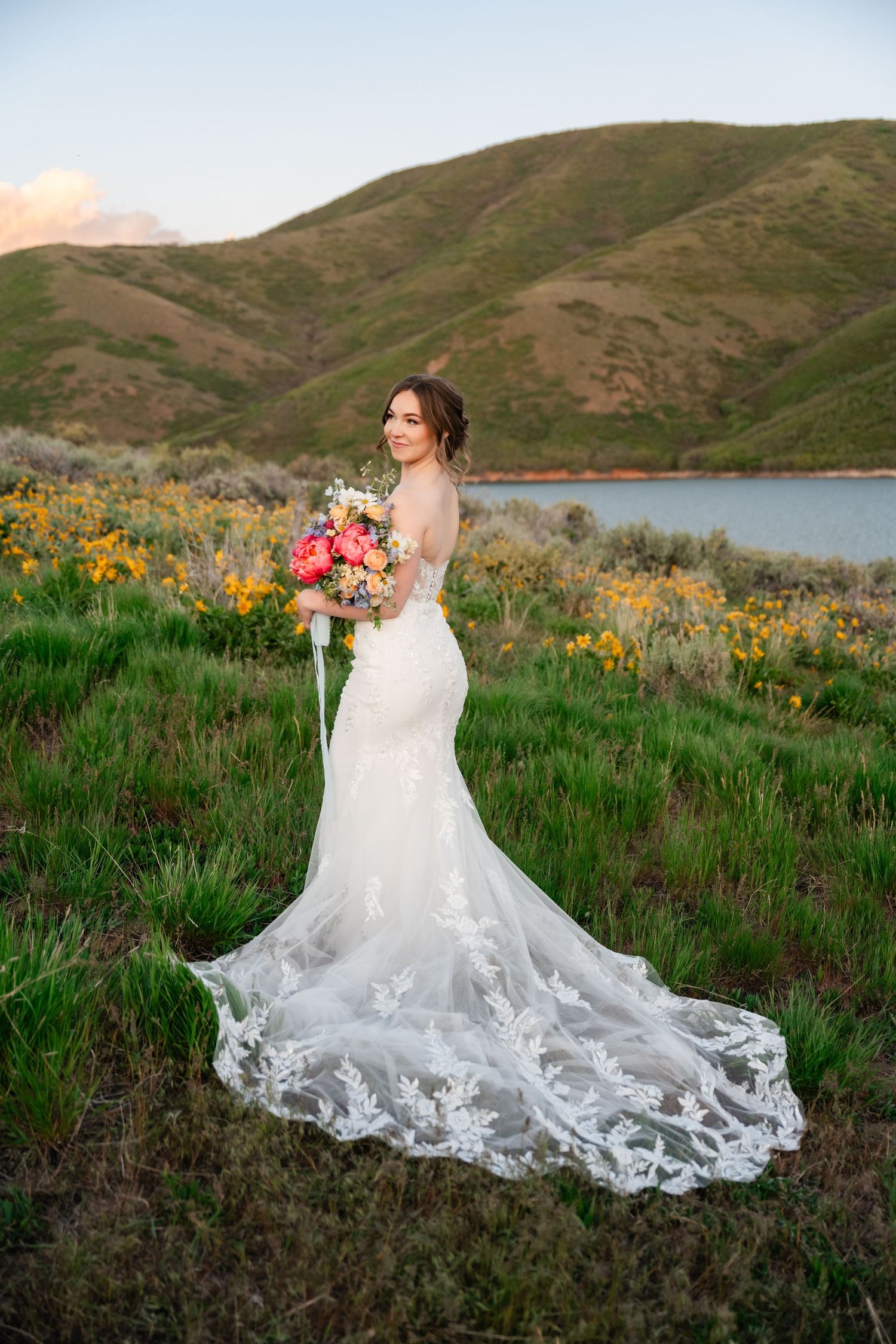 Bride's lace dress train flows beautifully across green grass with mountains and wildflowers in backdrop.