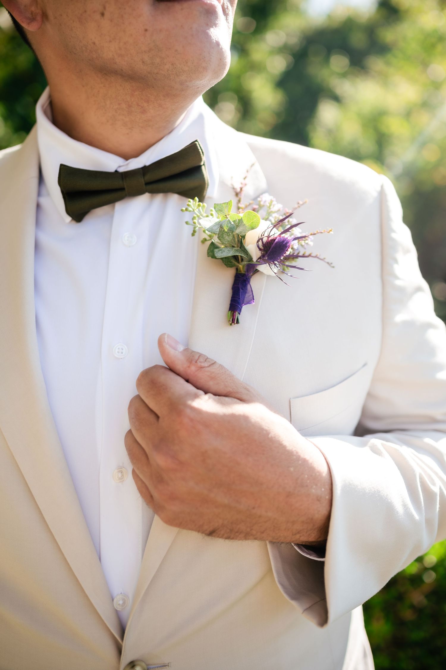Close up of a cream formal suit jacket with black bowtie and purple floral boutonniere at an outdoor wedding.