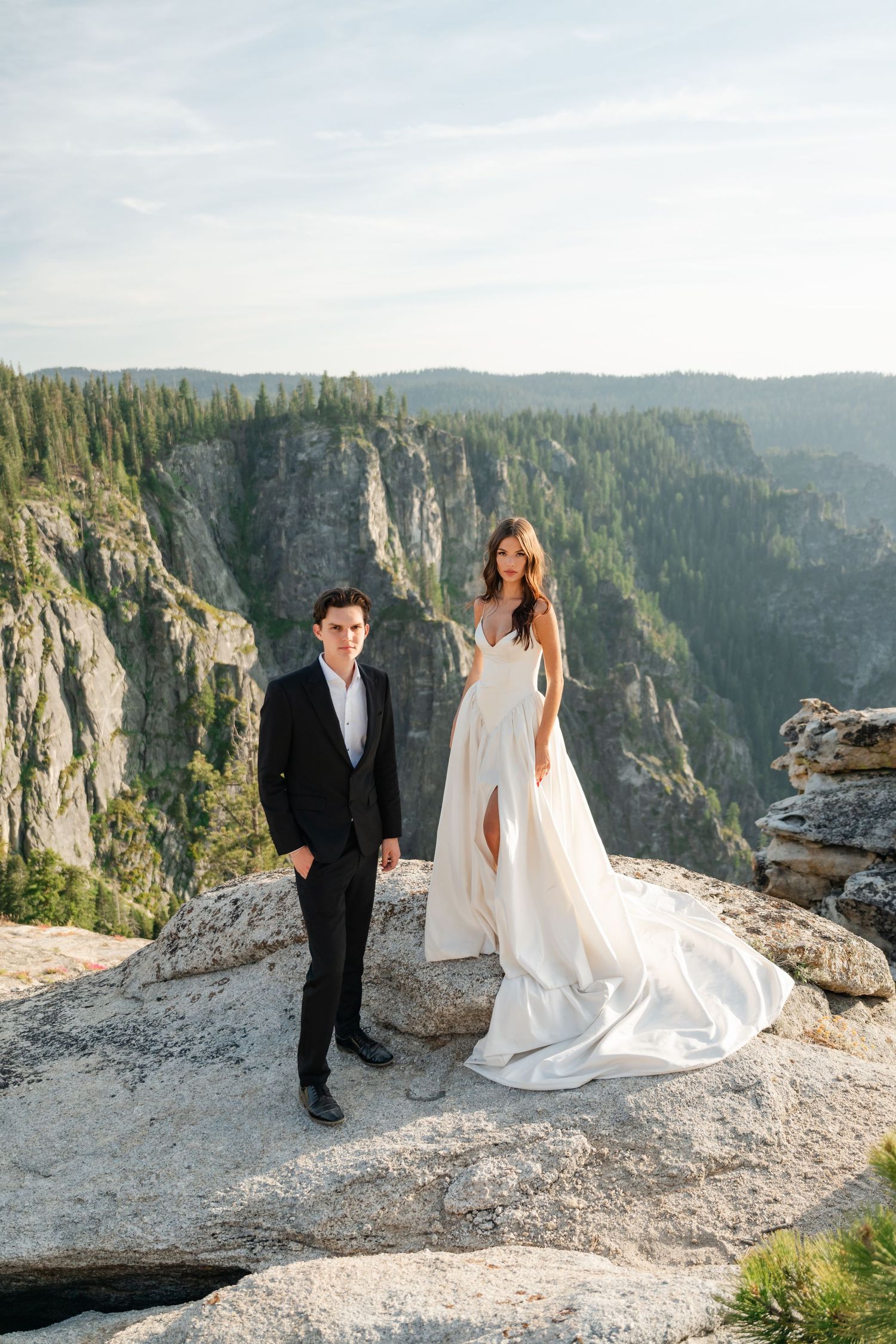 Bride and groom standing on rocky cliff edge with stunning mountain canyon backdrop during outdoor wedding photo session.