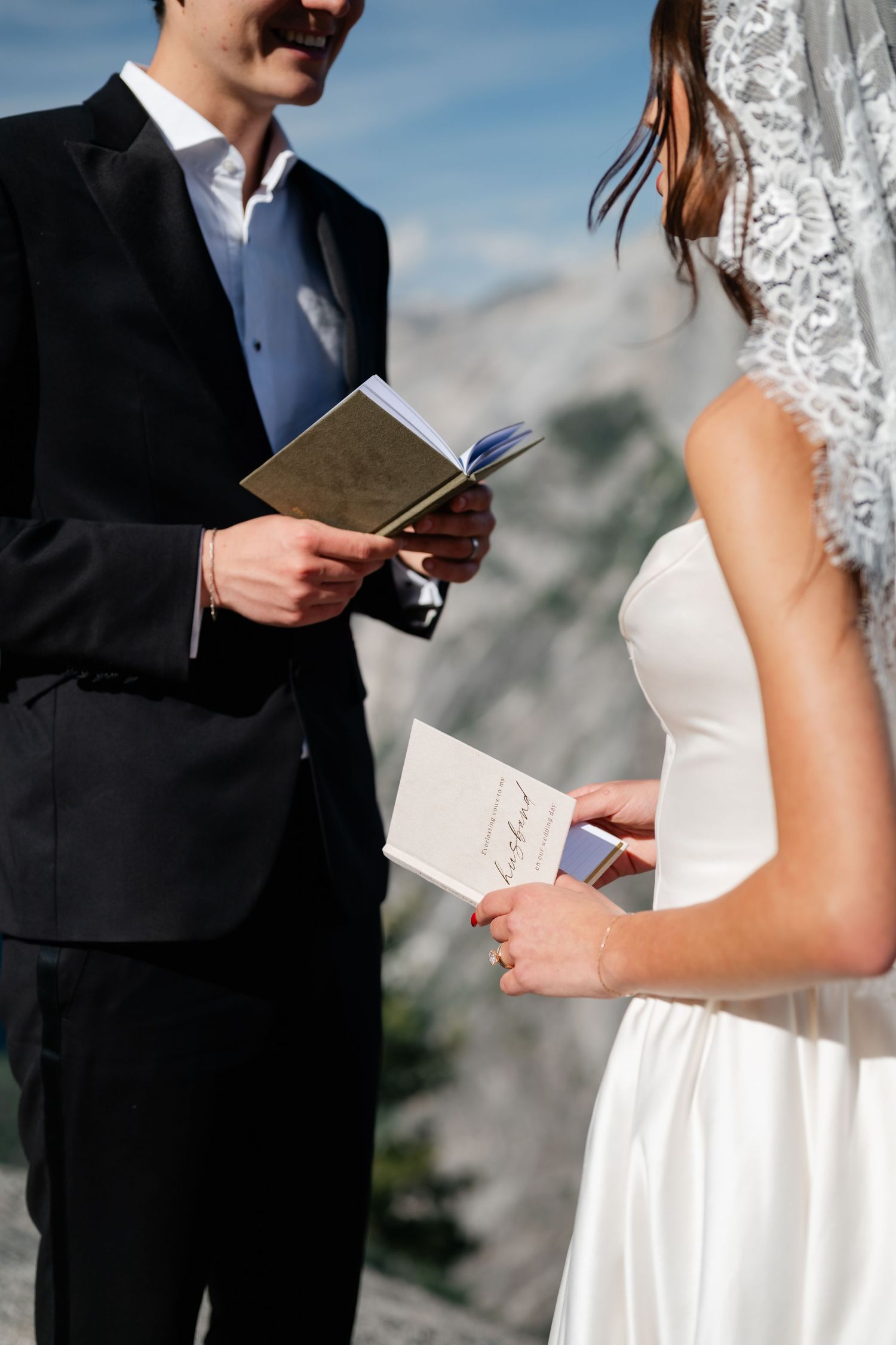 Bride and groom exchange vows holding booklets outdoors with mountain backdrop at elegant wedding ceremony.
