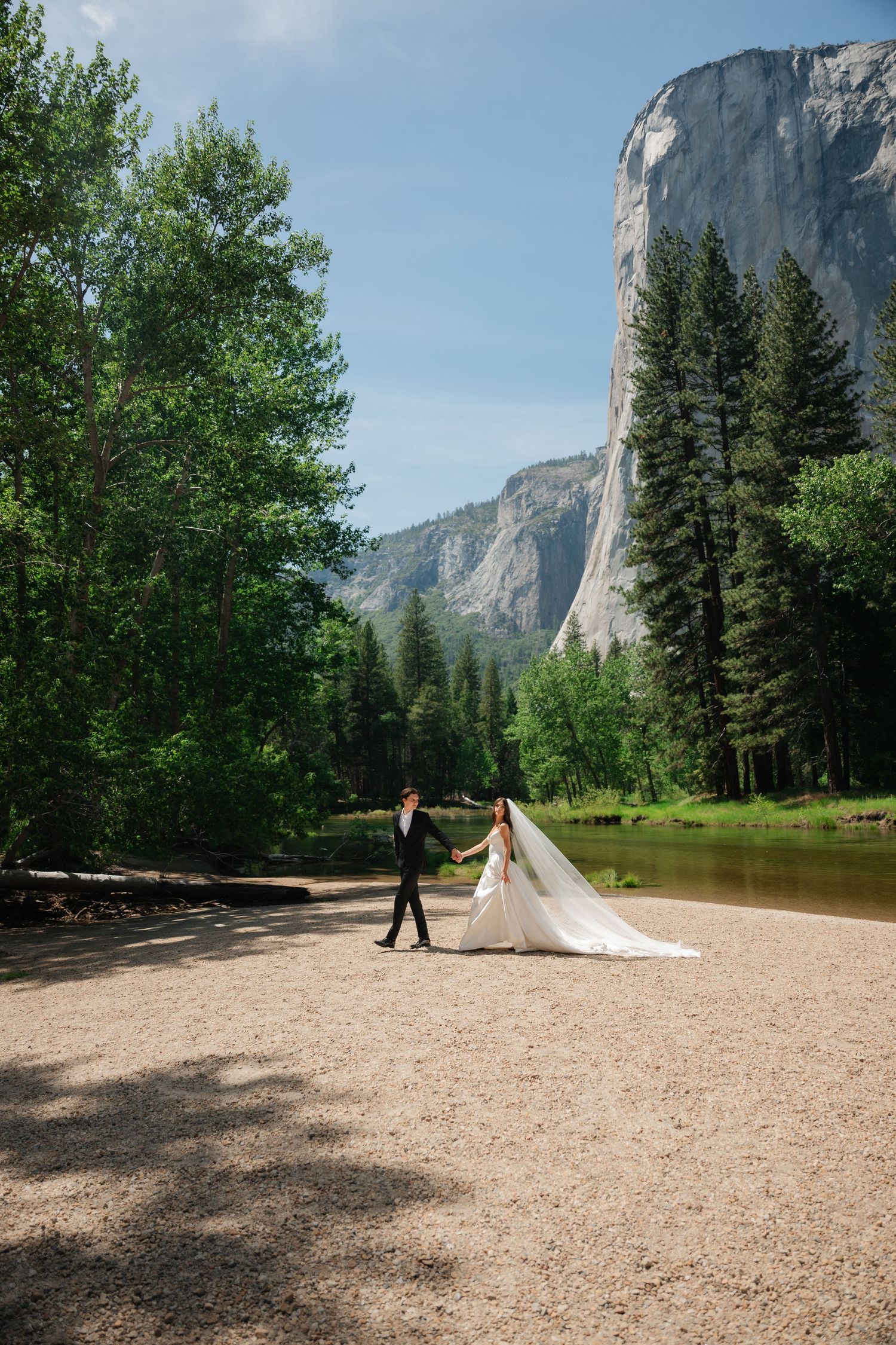 Bride and groom walk hand-in-hand along a sandy riverbank in Yosemite with El Capitan in the background.