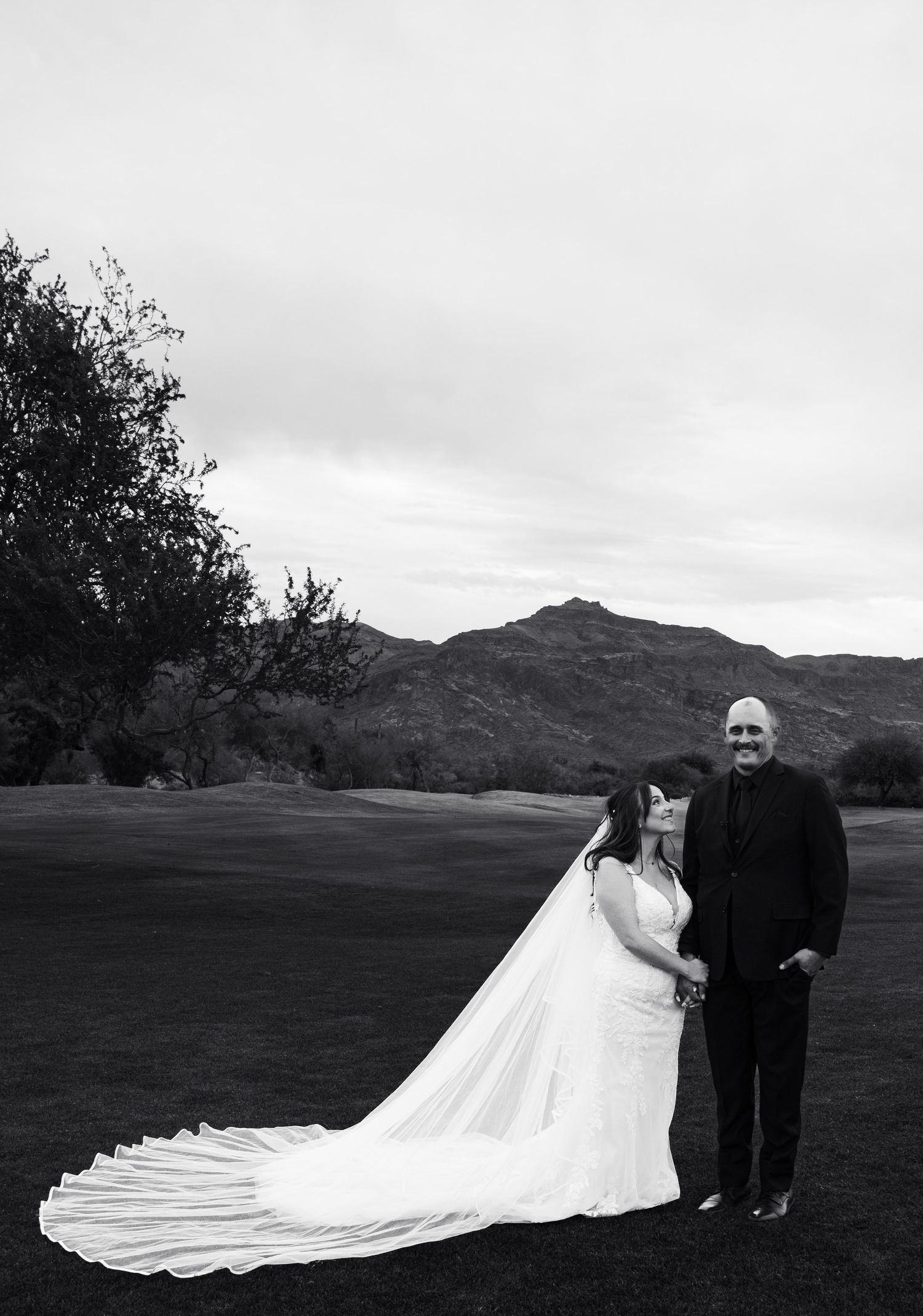 A wedding couple stands together on a golf course with mountains in the background as the bride's long veil flows behind them.