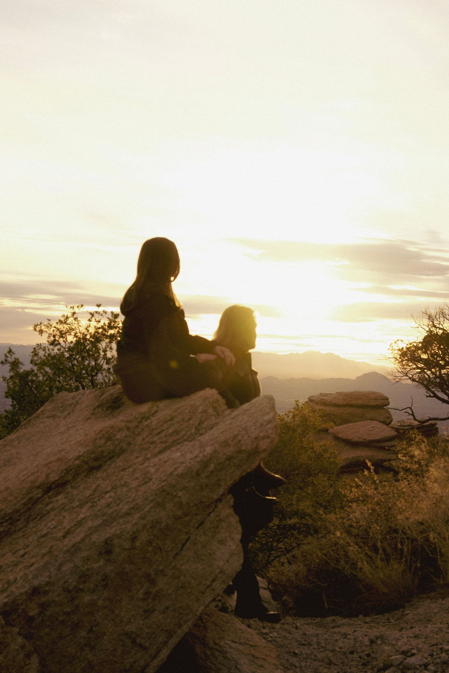 Two silhouettes sit together on a rocky outcrop during golden hour.