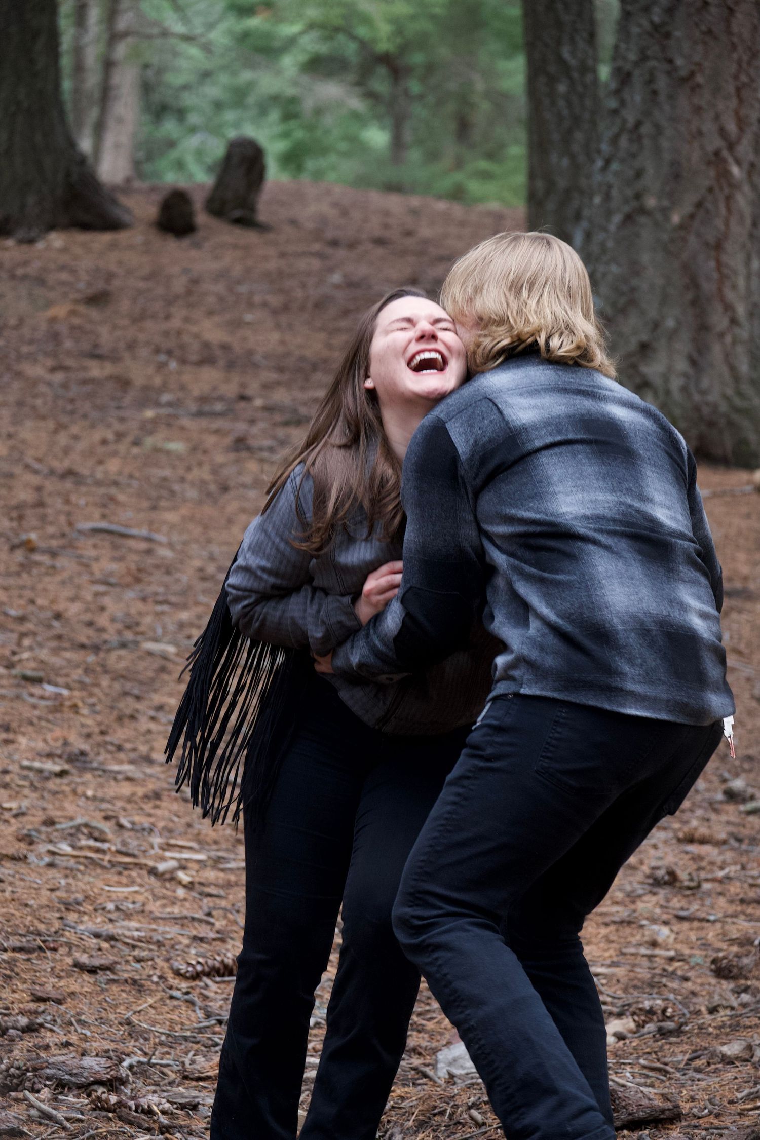 A couple laughing and playing in a forest setting wearing dark clothing.