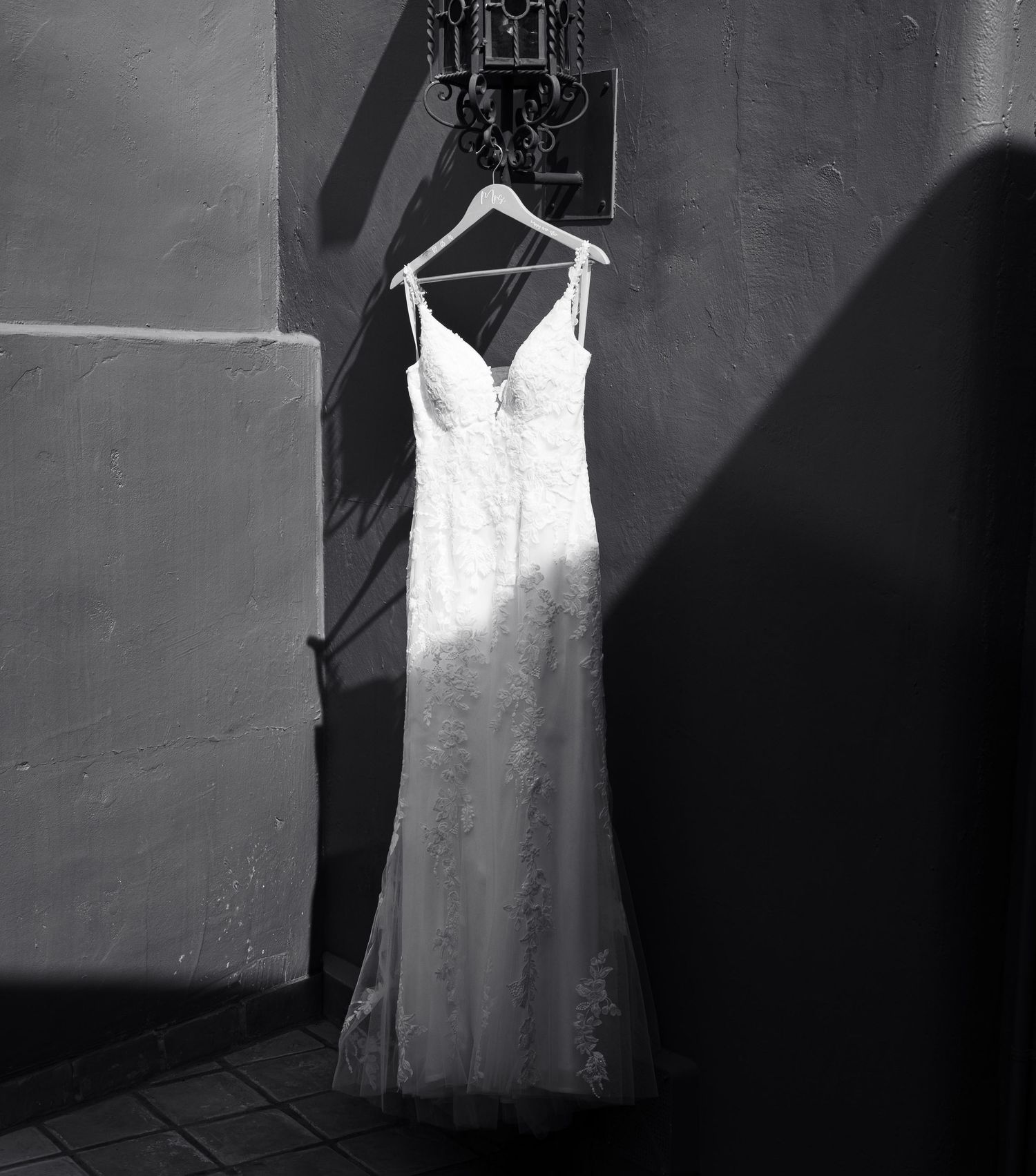 A white lace wedding dress hangs elegantly on a hanger against a shadowed wall in dramatic black and white lighting.