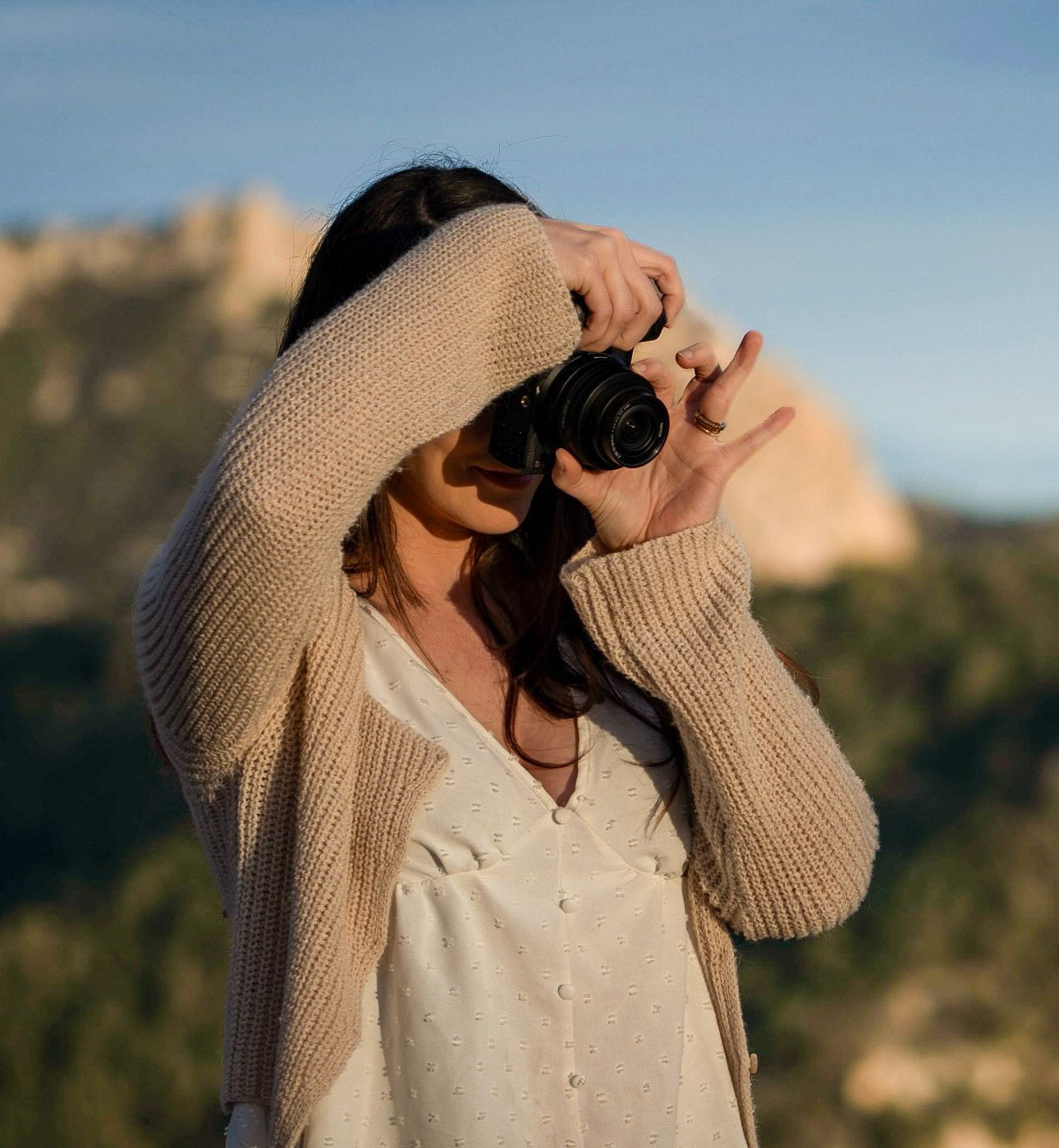 A photographer takes photos in golden sunlight while wearing a cream colored sweater.