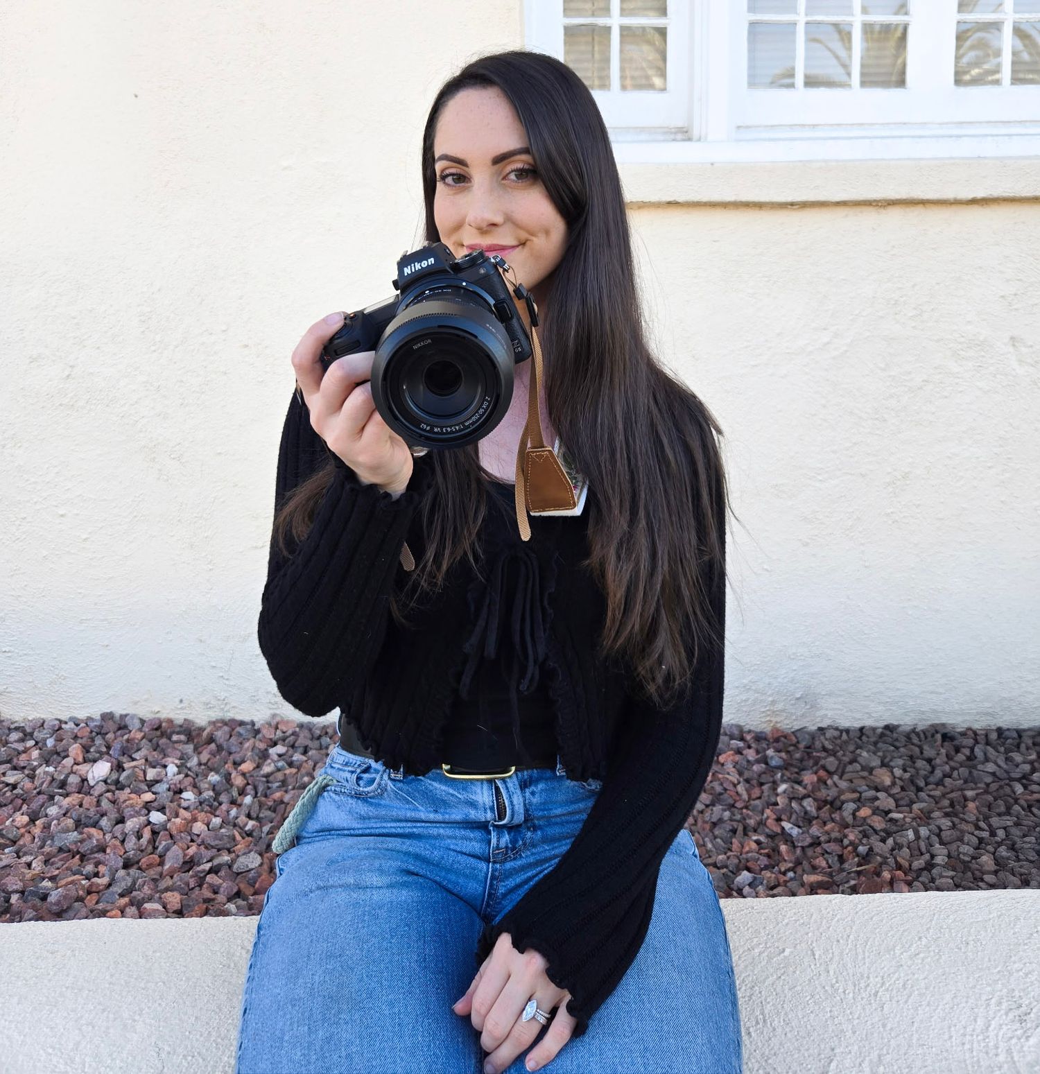 A photographer in a black sweater and blue jeans sits casually while holding a professional camera outdoors.