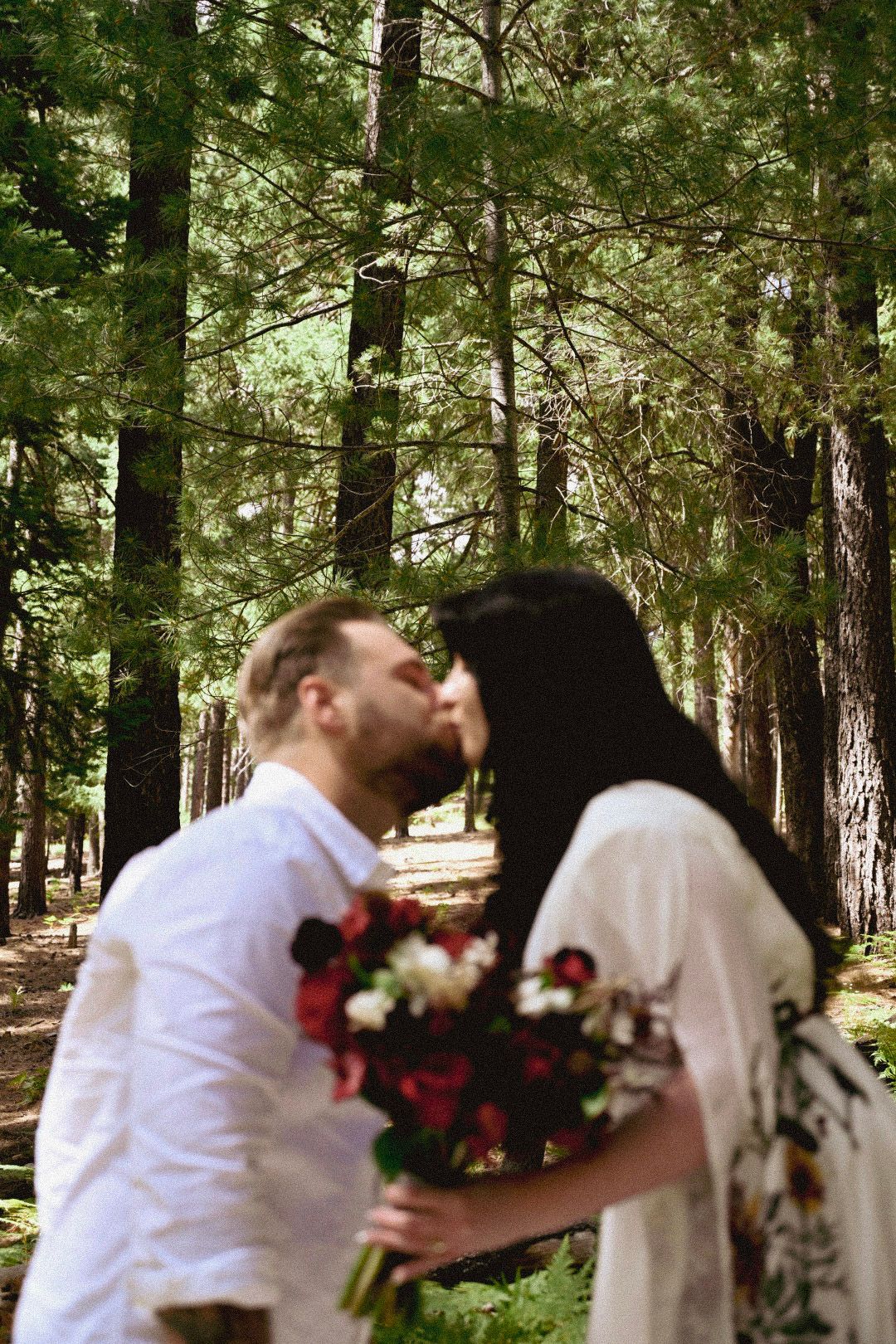 A romantic forest scene with red floral bouquet and white attire among tall pine trees.