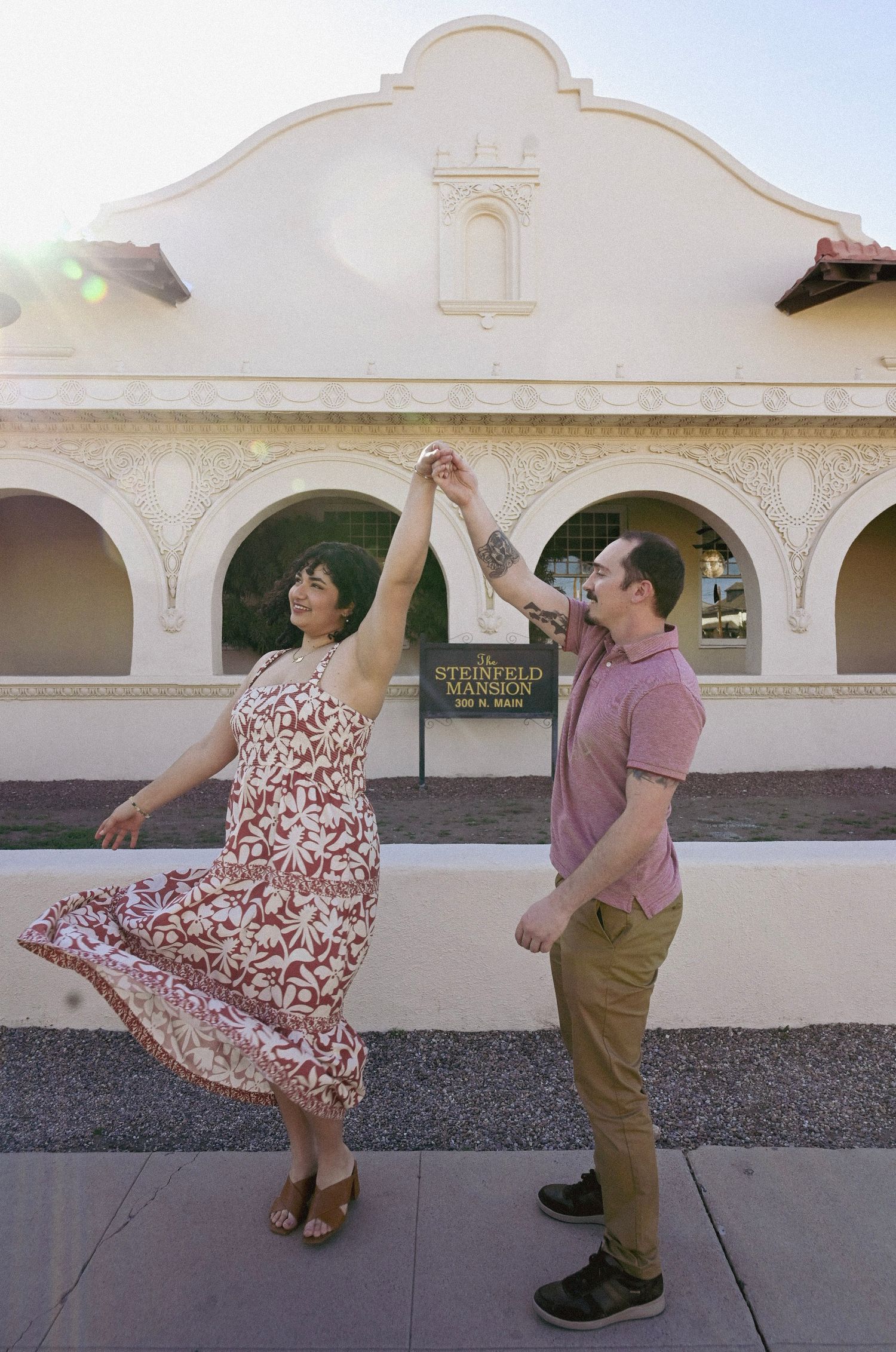 A couple dances playfully in front of a Spanish colonial style building with arched windows and decorative facade.