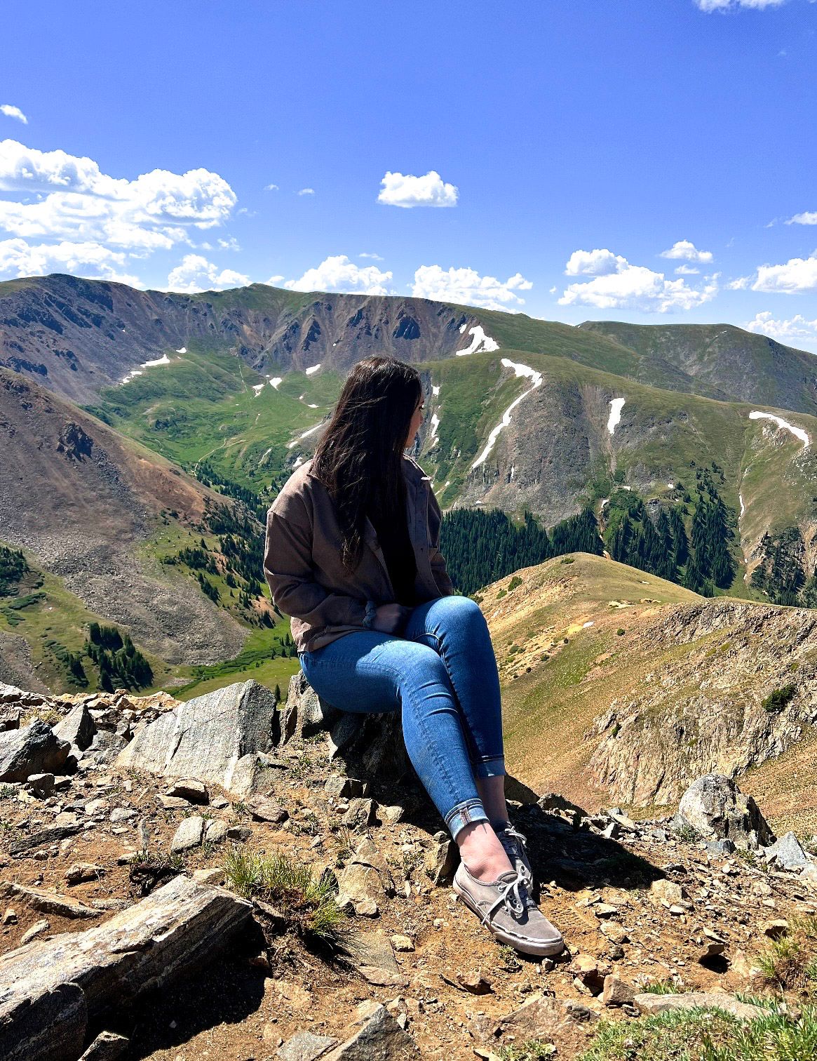 A person in blue jeans lies on their stomach at the edge of a rocky mountain overlook with scenic mountain views.