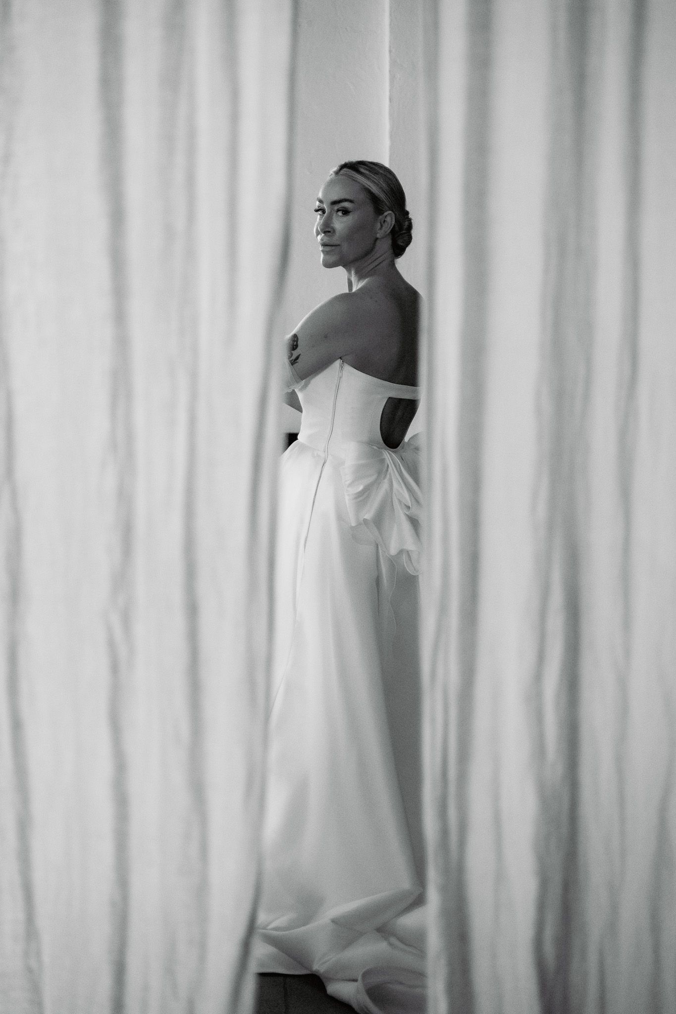 Editorial stijl zwart-witfoto van een bruid in een strapless jurk die tussen witte gordijnen staat.