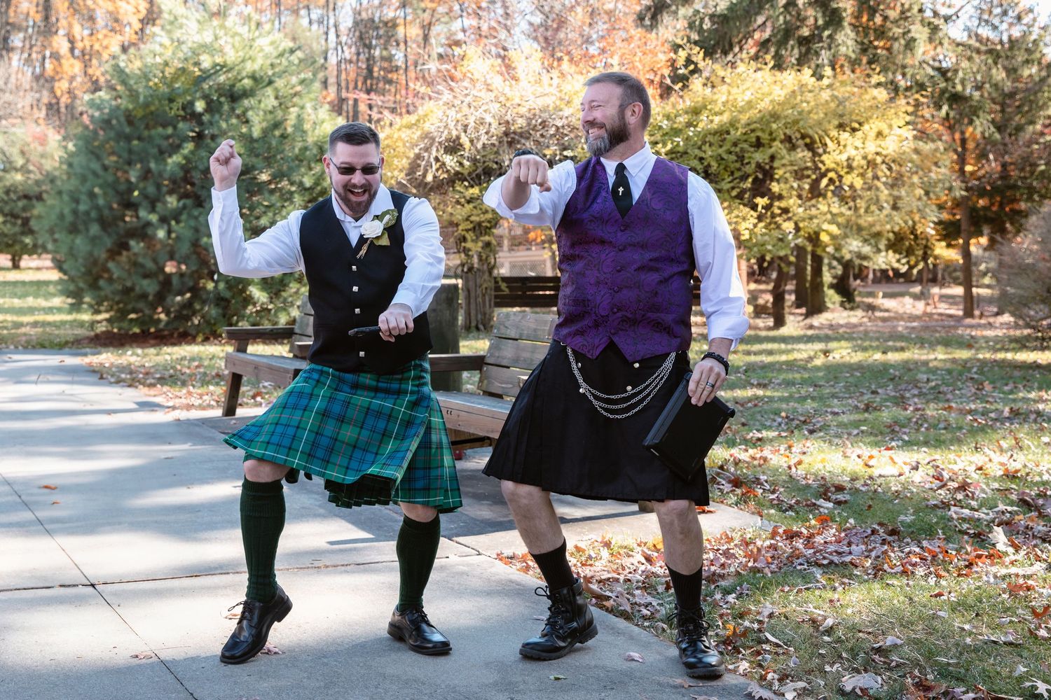Two people in kilts pose playfully together outdoors in an autumn setting.
