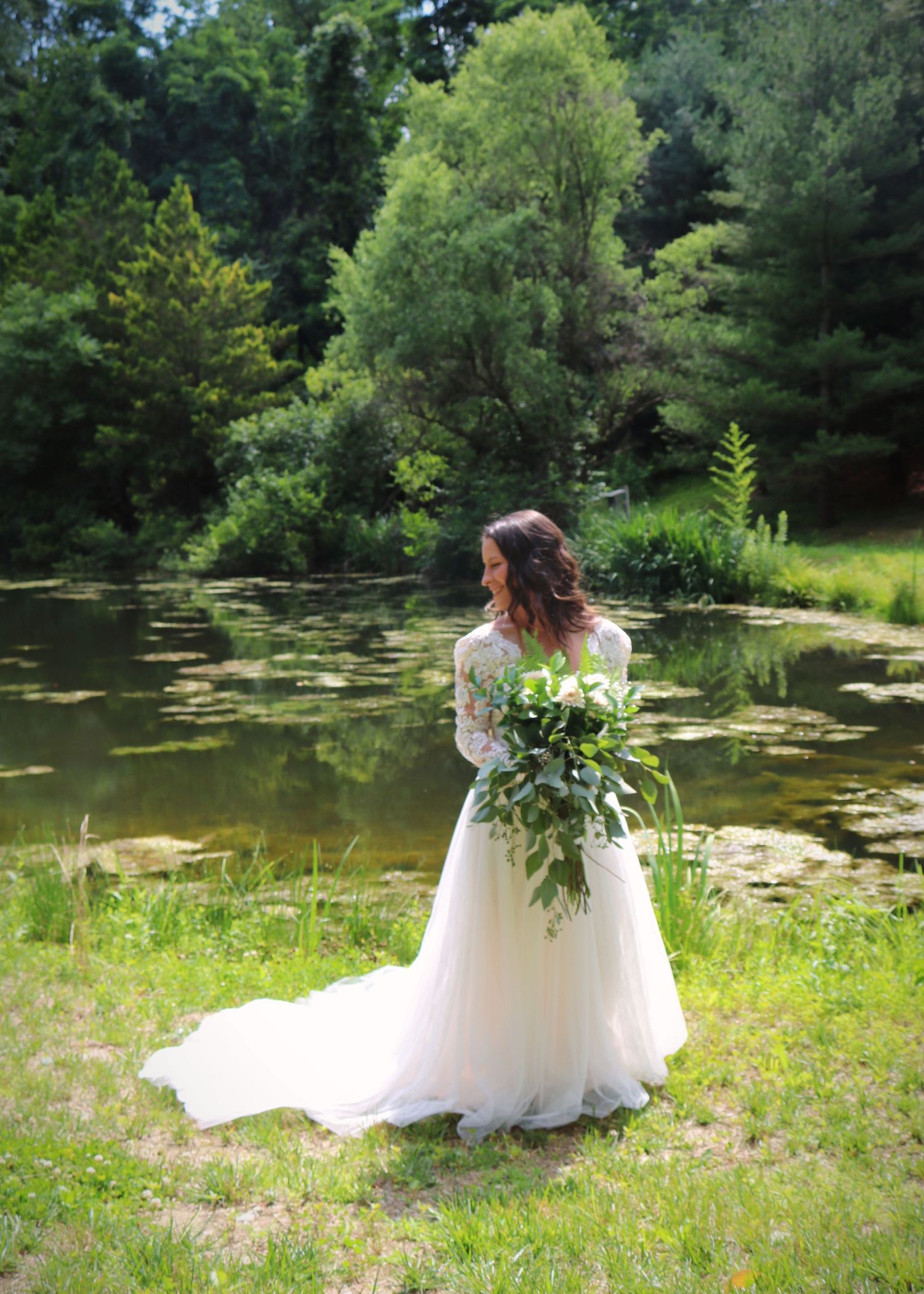 Bride in white gown holding greenery bouquet by pond in garden setting.