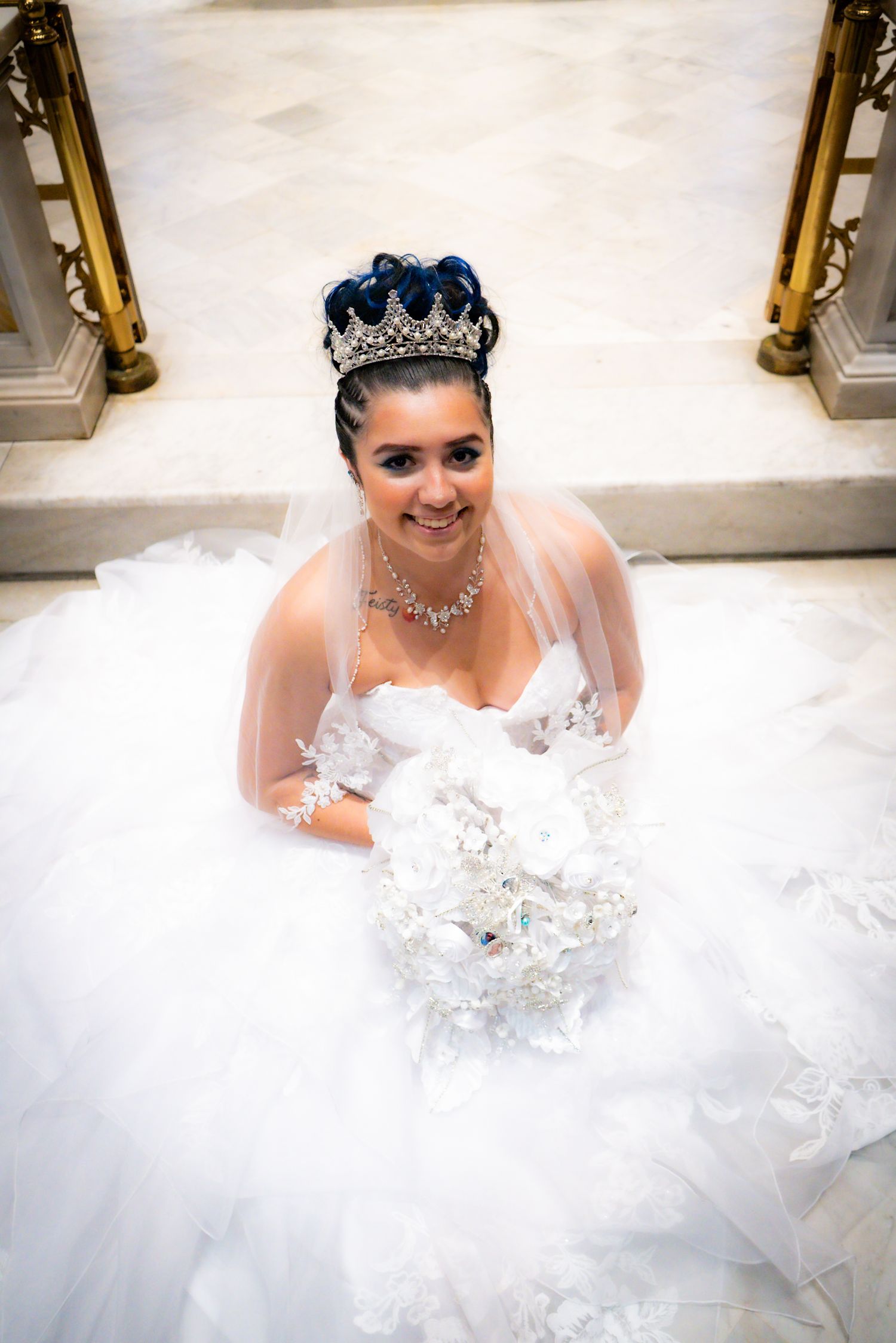 A bride in a white gown and blue crown sits on a marble floor.