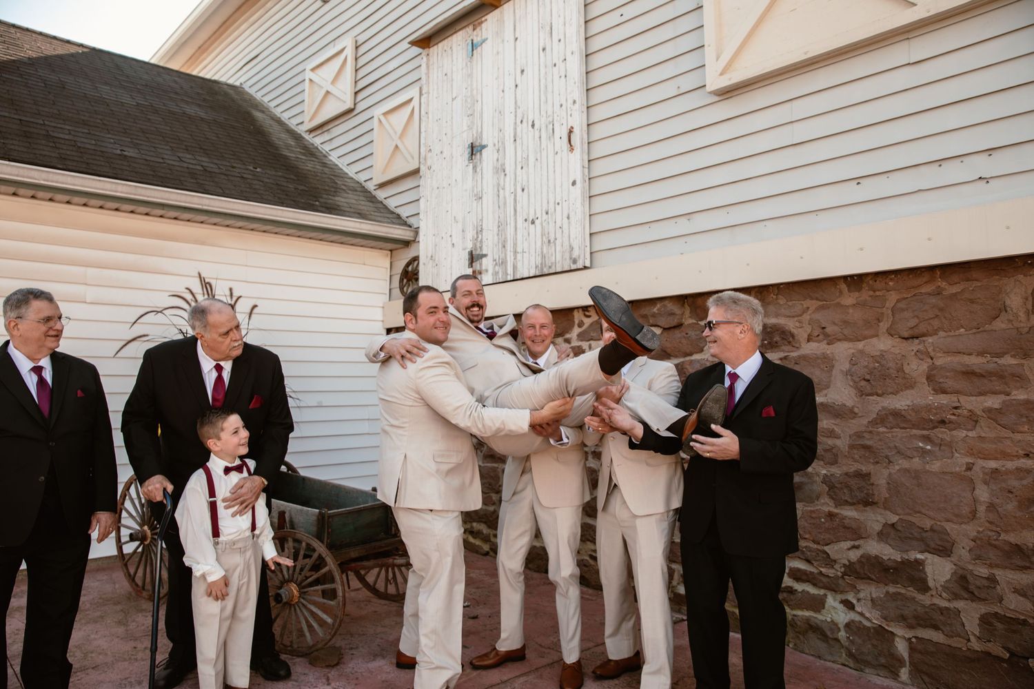 Wedding party in light and dark attire poses together against white clapboard siding and stone walls.