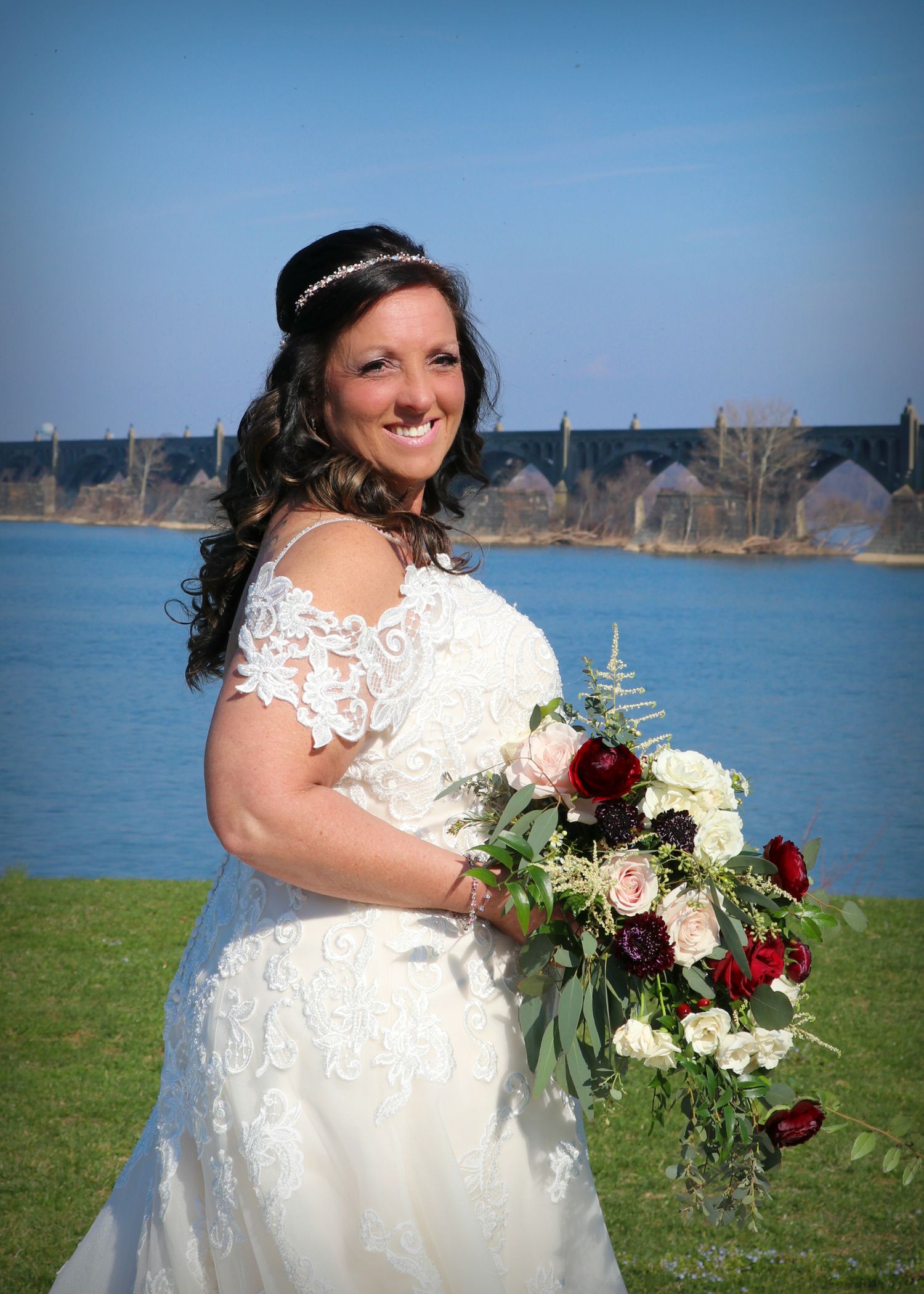 Bride in lace dress with burgundy bouquet poses by waterfront on sunny day.