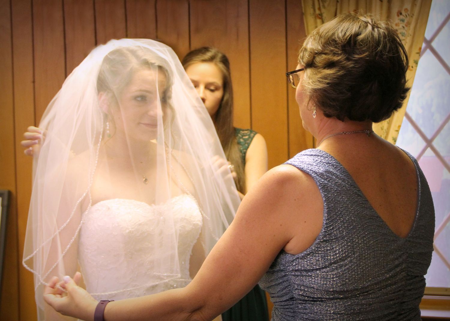 Final adjustments being made to a bride's white veil in a wood-paneled room.