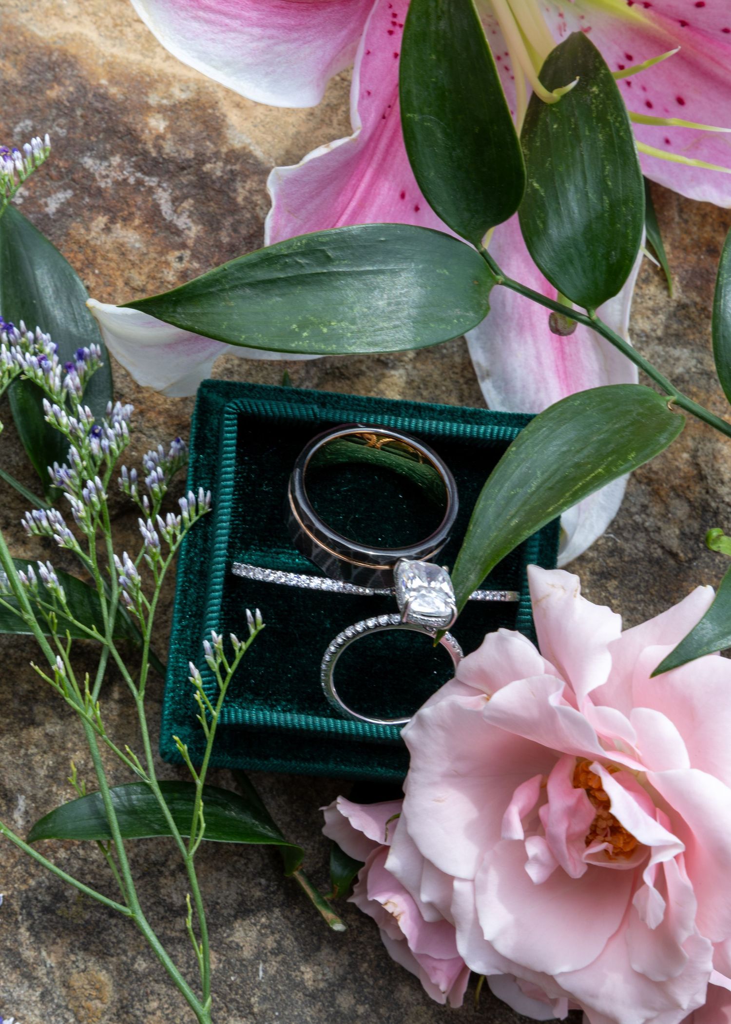 Wedding rings nestled in a dark green velvet box with pink lilies, roses and delicate purple flowers on stone surface.