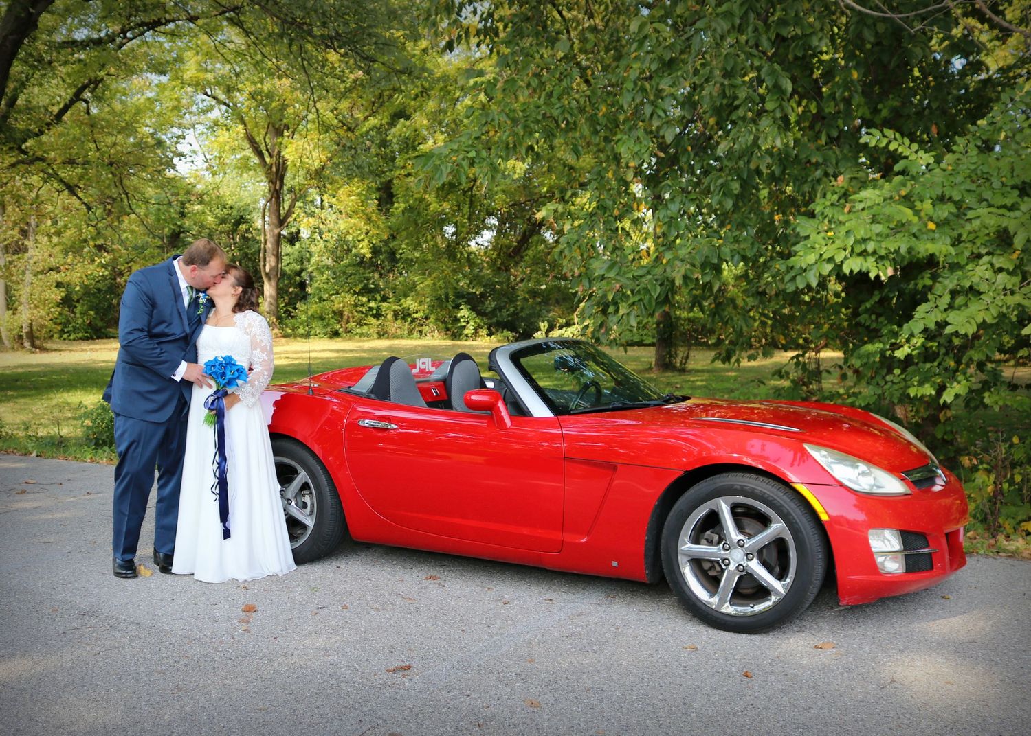 Newlywed couple sharing an intimate moment next to a red sports car.