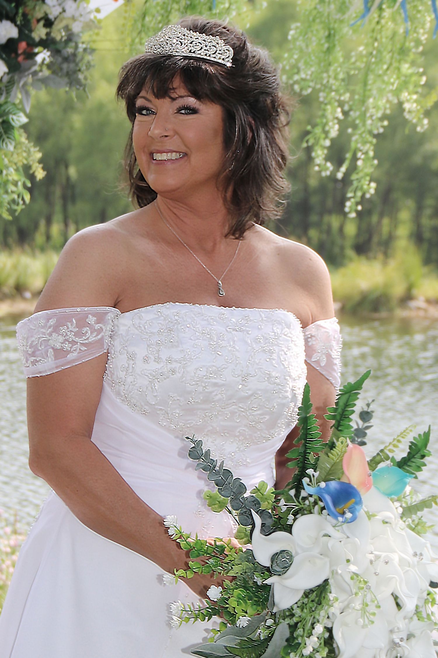 Bride in off-shoulder dress poses with bouquet against willow tree background.