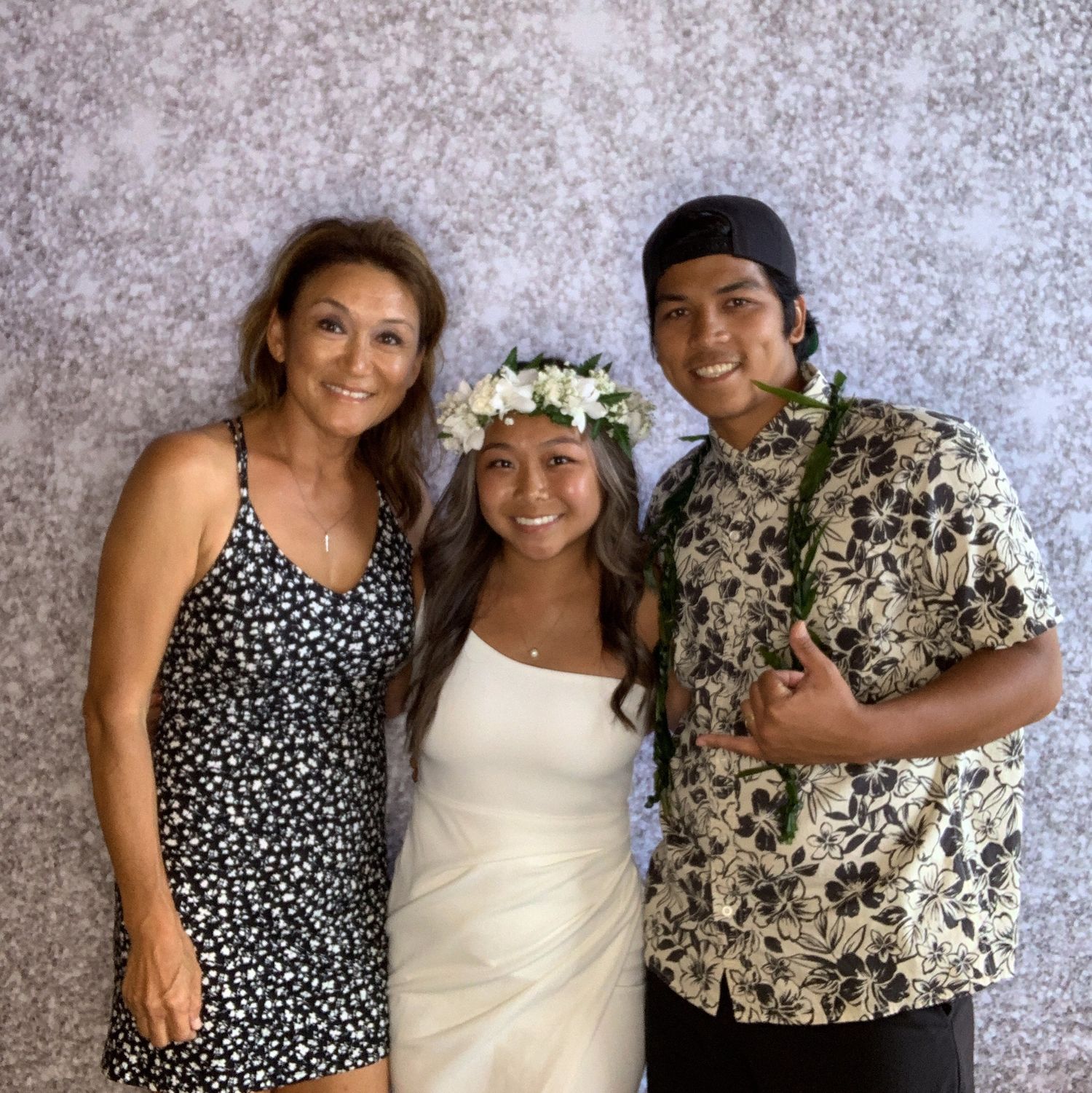 Three people in casual attire posing together, with one wearing a floral crown and white dress.