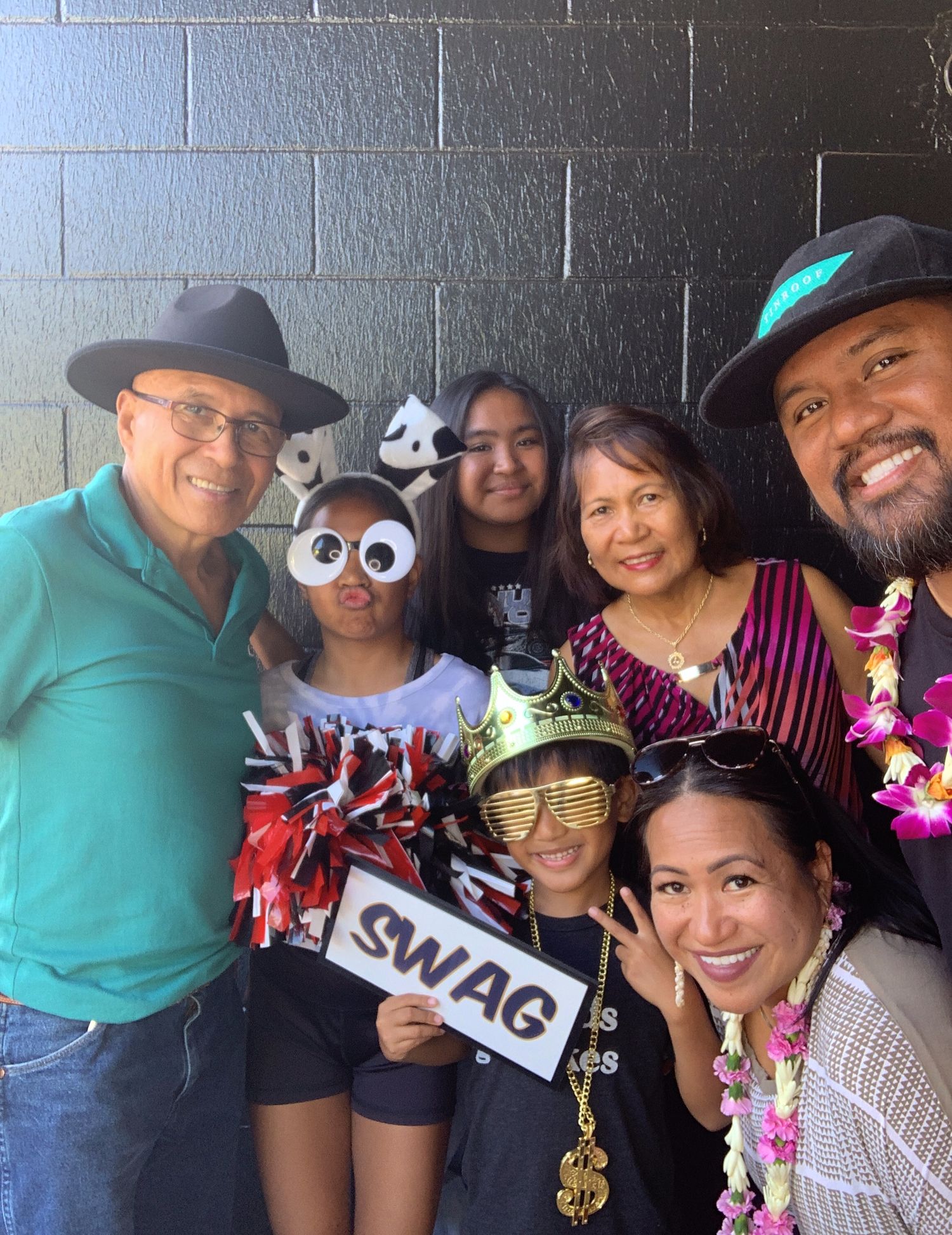 Group photo with fun props including googly eye glasses, a SWAG sign, and tropical leis against a brick wall background.