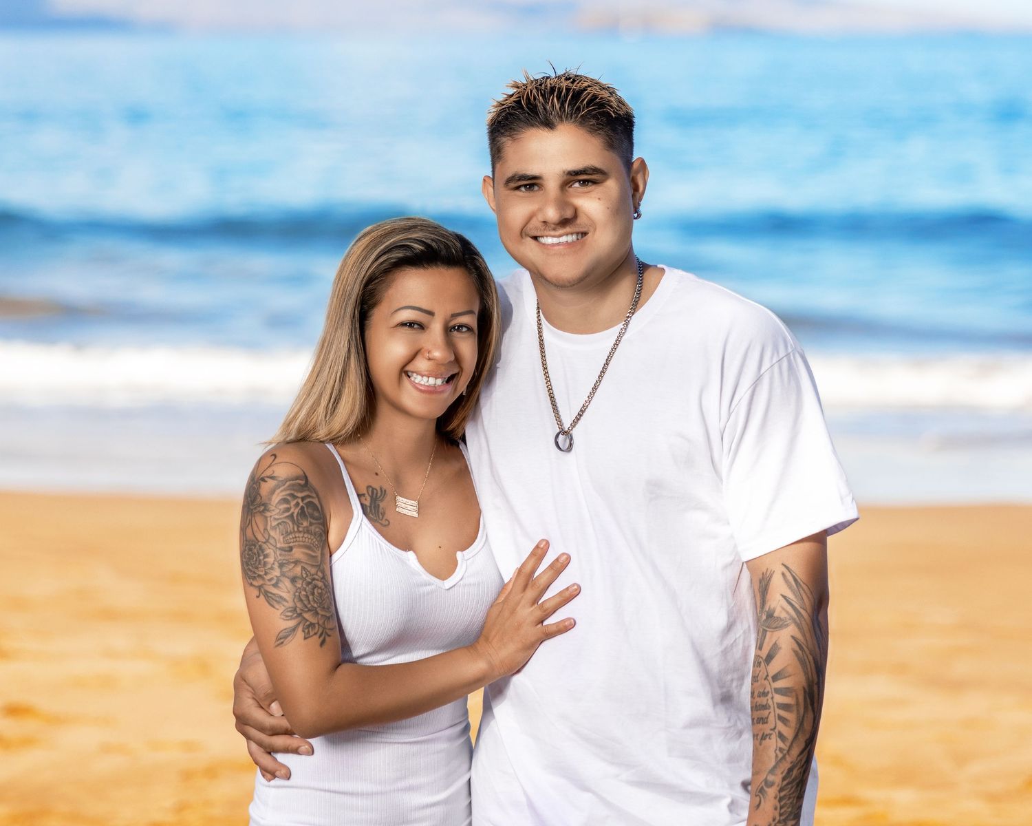 Couple in white clothing embrace on beach with blue ocean and sky behind them.