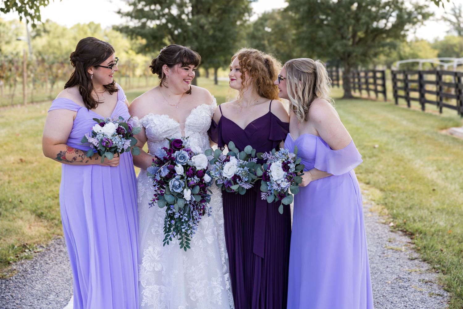 A wedding party in coordinated purple and lavender dresses holds bouquets while standing together outdoors.