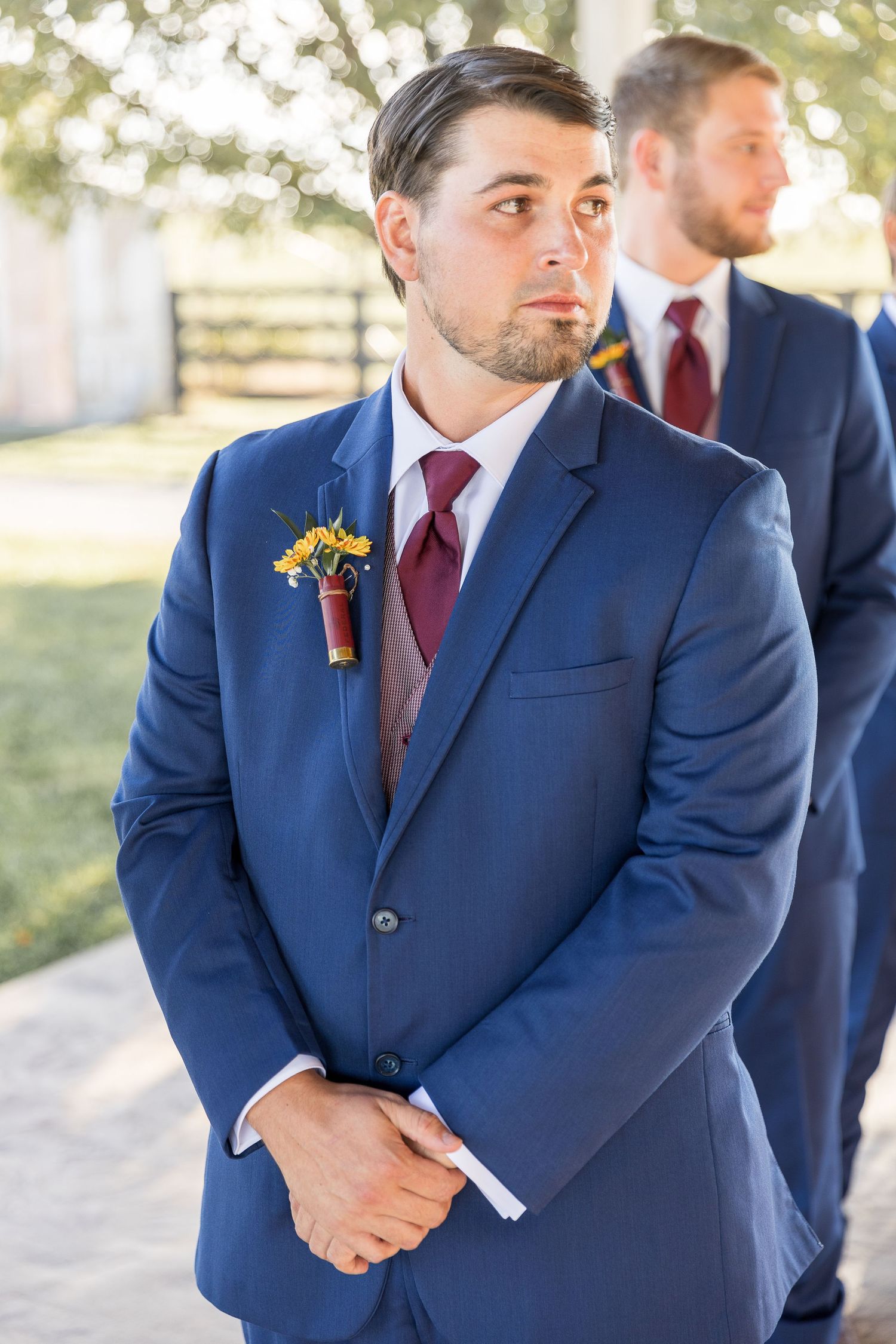 Groomsman in navy suit with burgundy tie and sunflower boutonniere at outdoor wedding.
