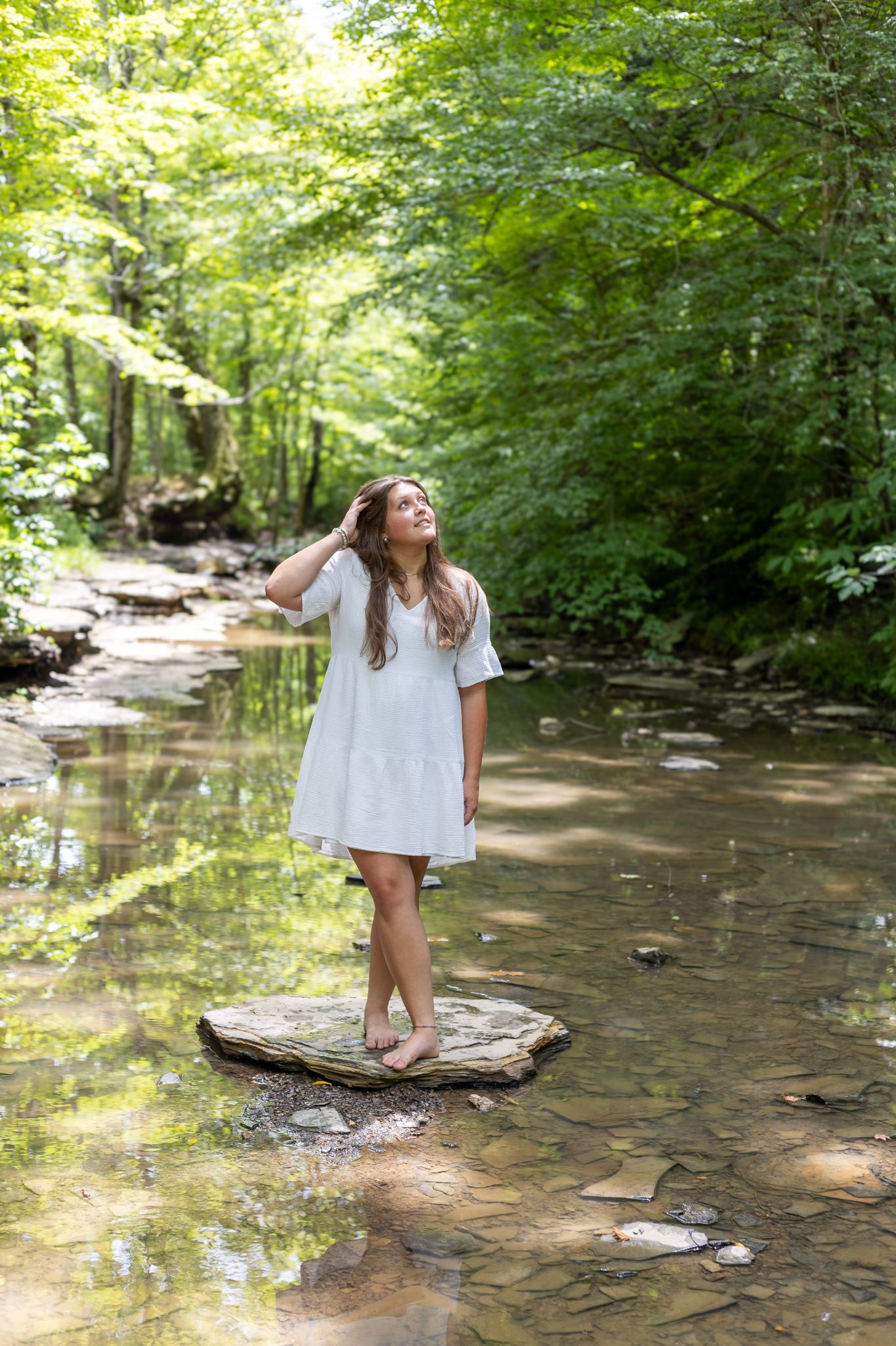 A person in a white dress stands on rocks in a shallow creek surrounded by lush green forest.