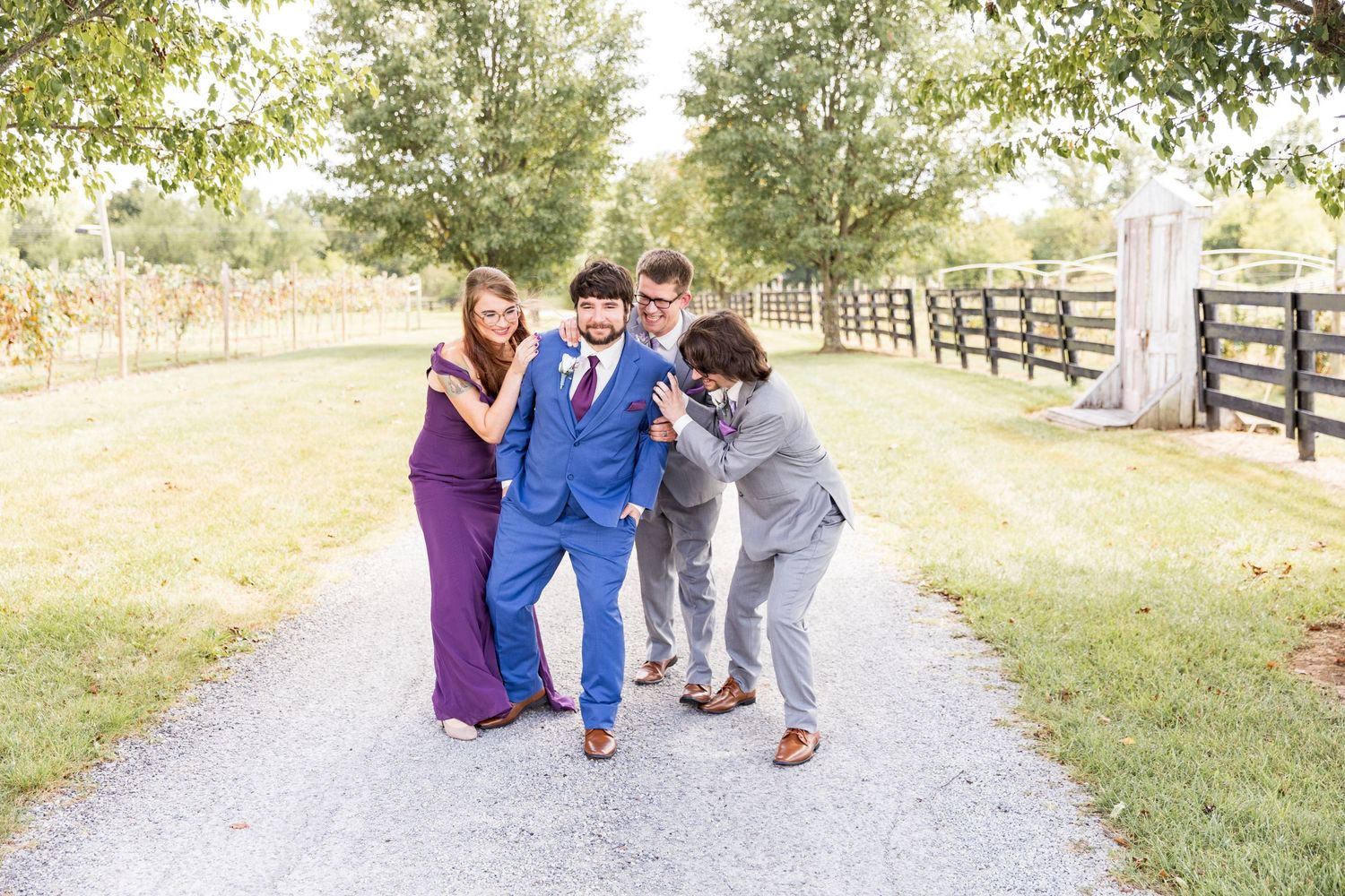 Friends in formal attire playfully support each other while walking down a country path.