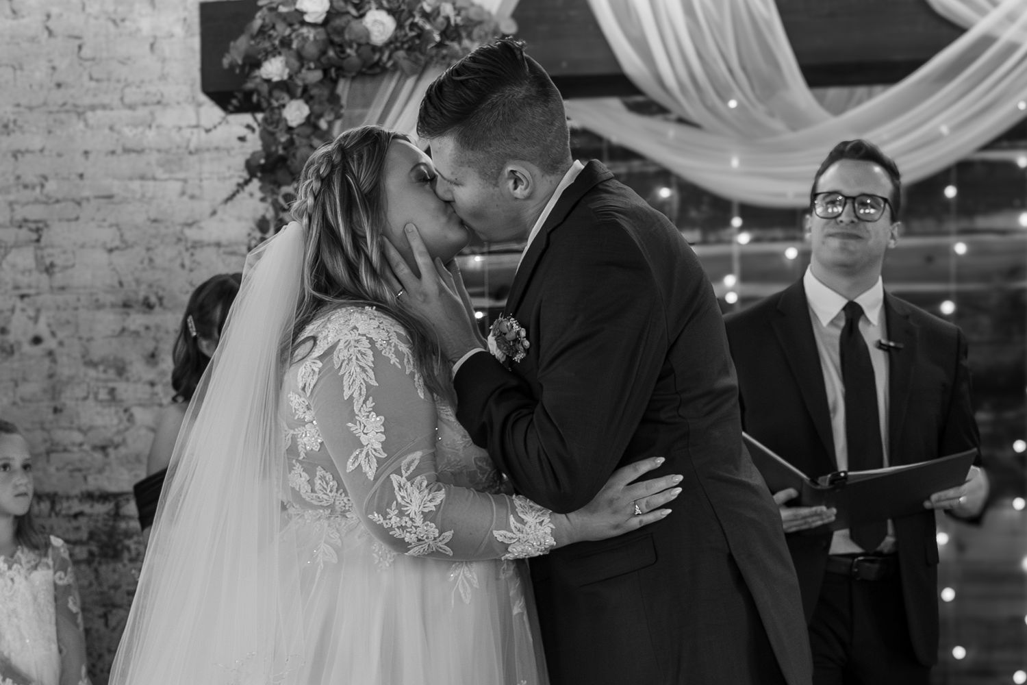A couple shares their first kiss during an indoor wedding ceremony with twinkling lights.