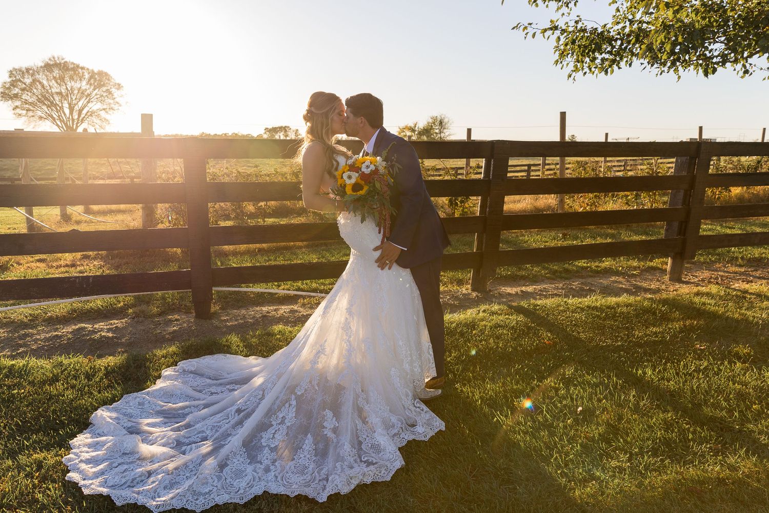 A couple shares a romantic moment at sunset by a wooden fence in their wedding attire.