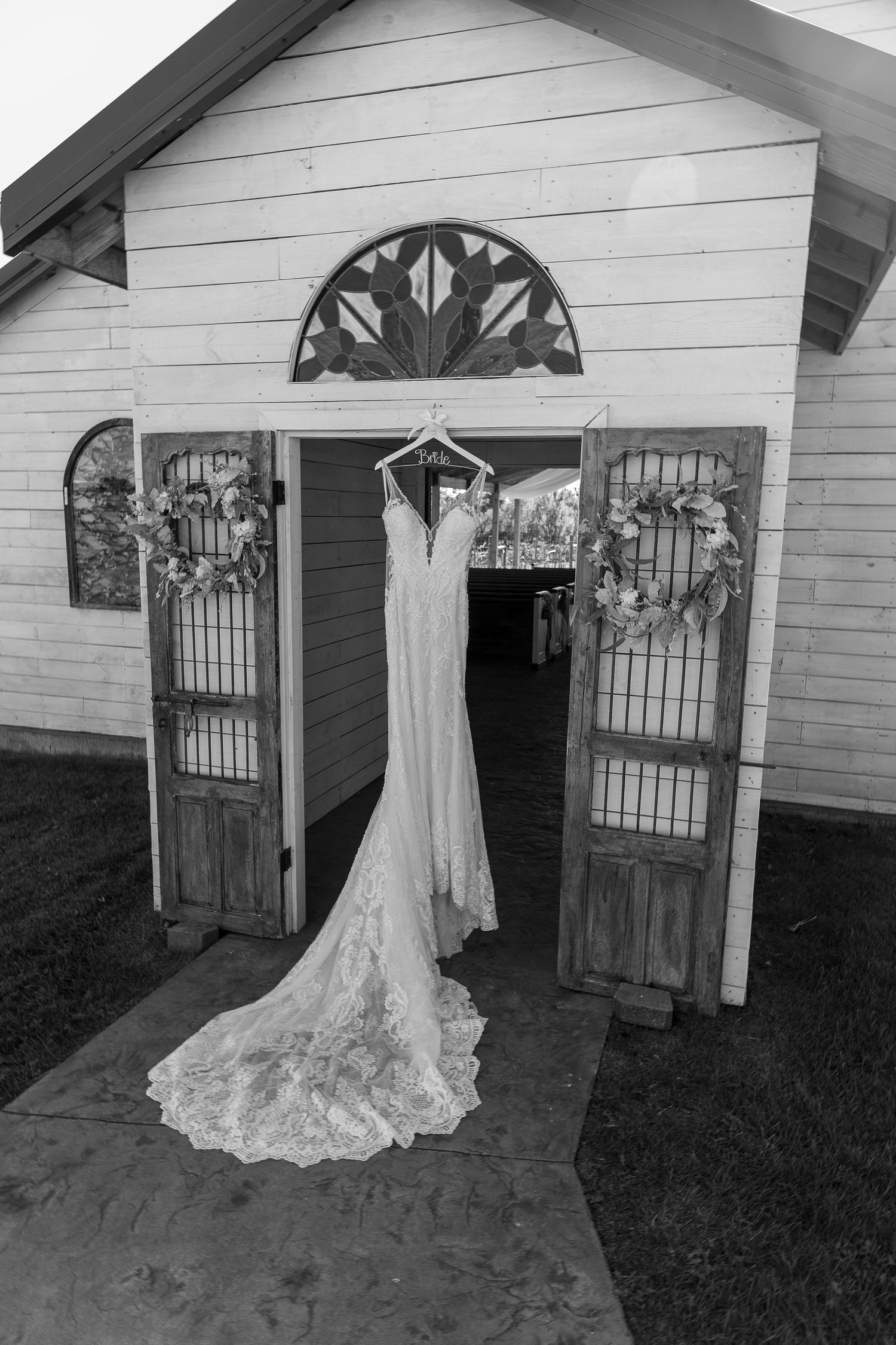 Elegant lace wedding dress hanging on rustic white chapel doors in black and white.