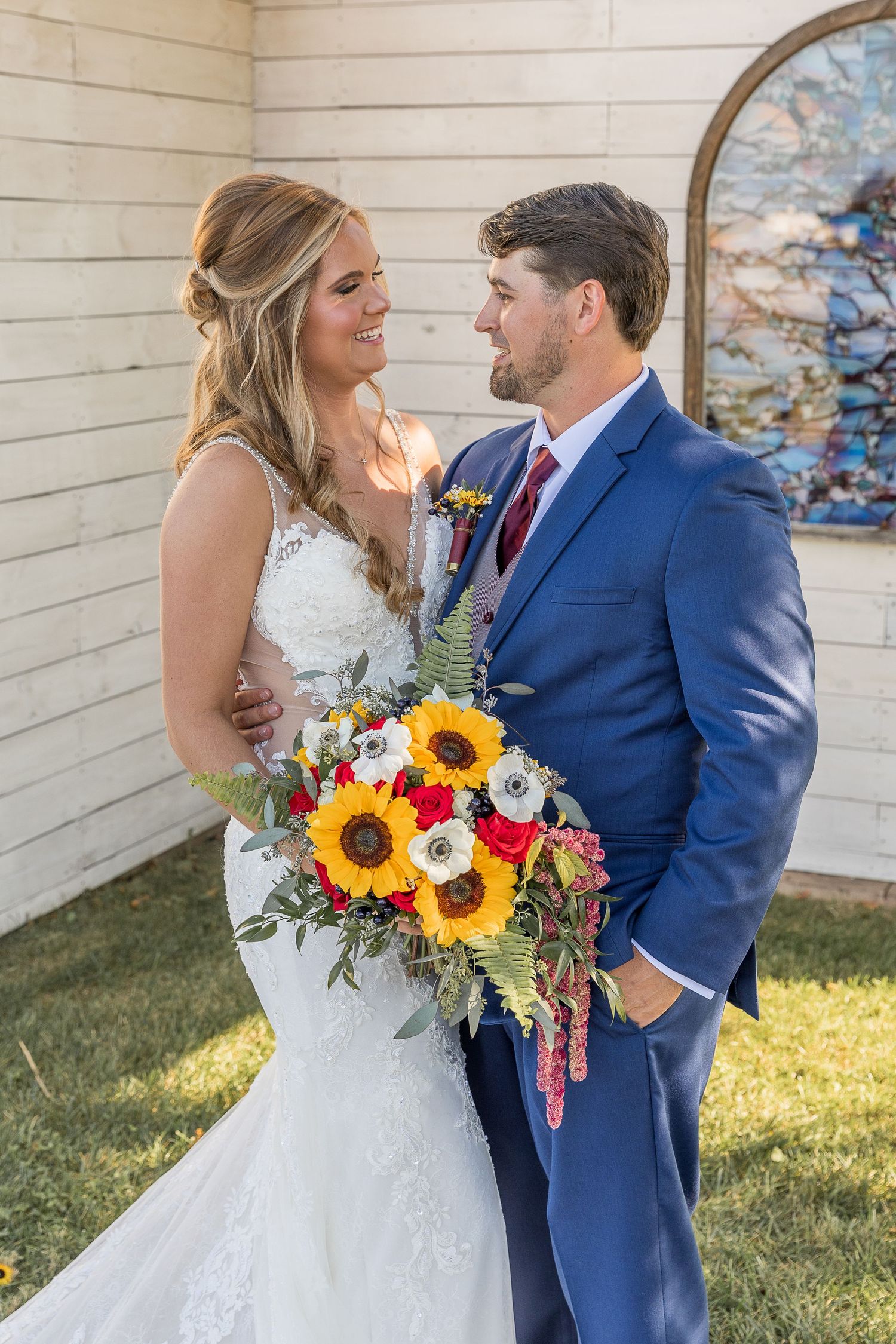 A wedding couple poses with vibrant sunflower bouquet against white wooden walls.