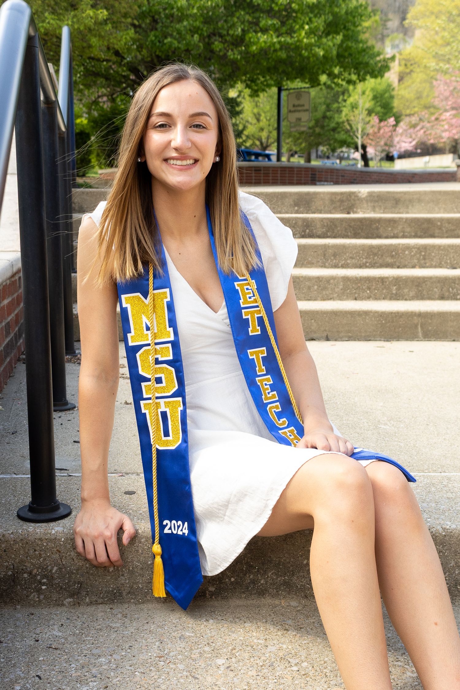 Graduate in white dress with blue and gold university sash sitting on concrete steps.