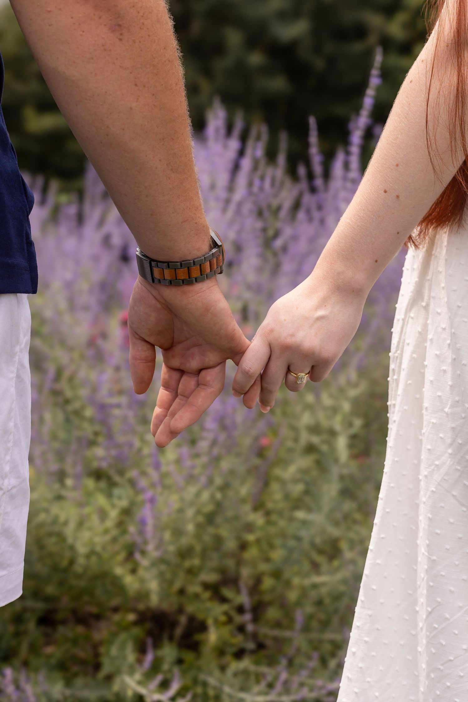 Close-up of couple holding hands with engagement rings visible against purple wildflowers.