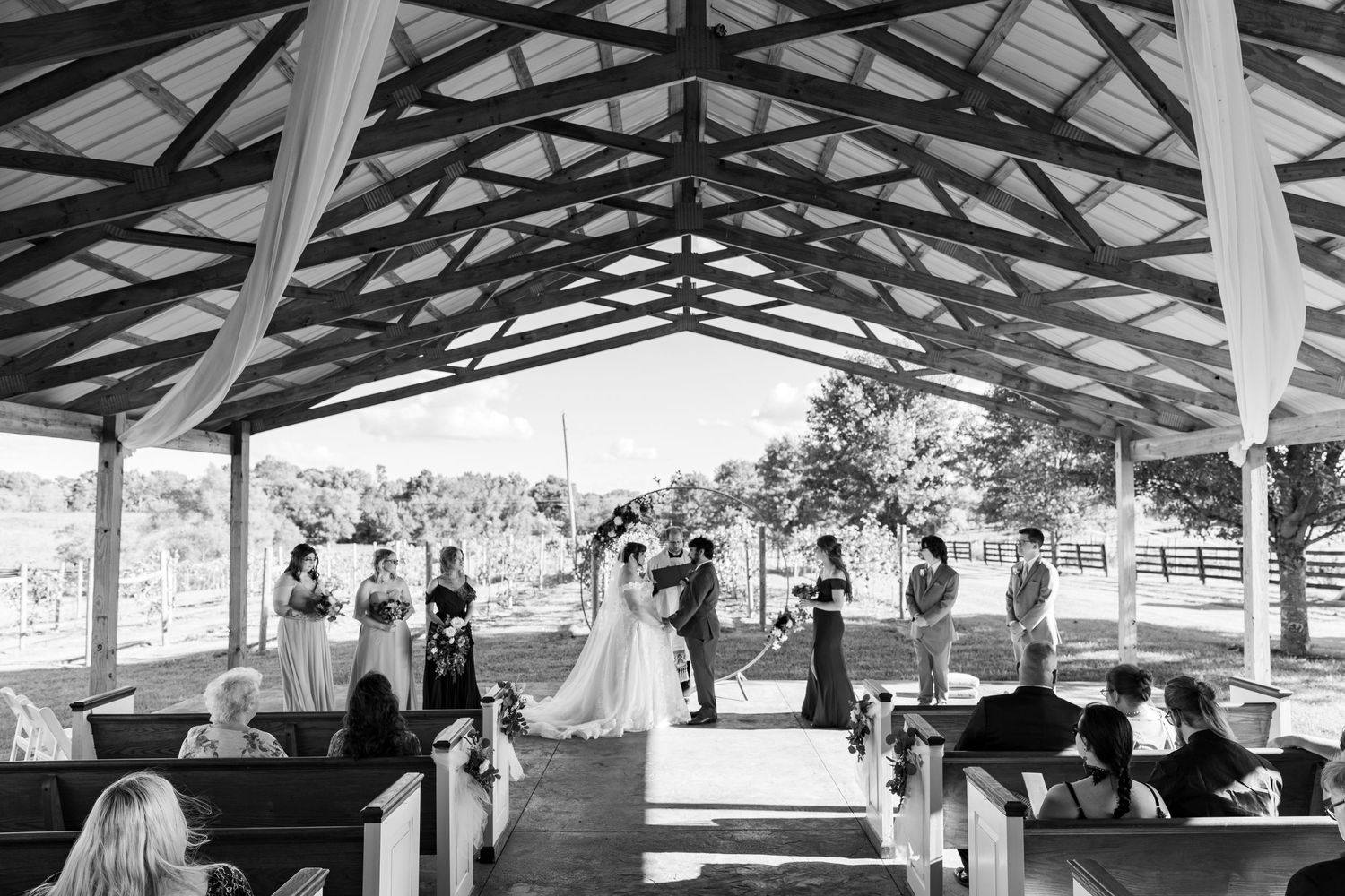 A wedding ceremony takes place under a rustic wooden pavilion with guests seated on wooden pews.