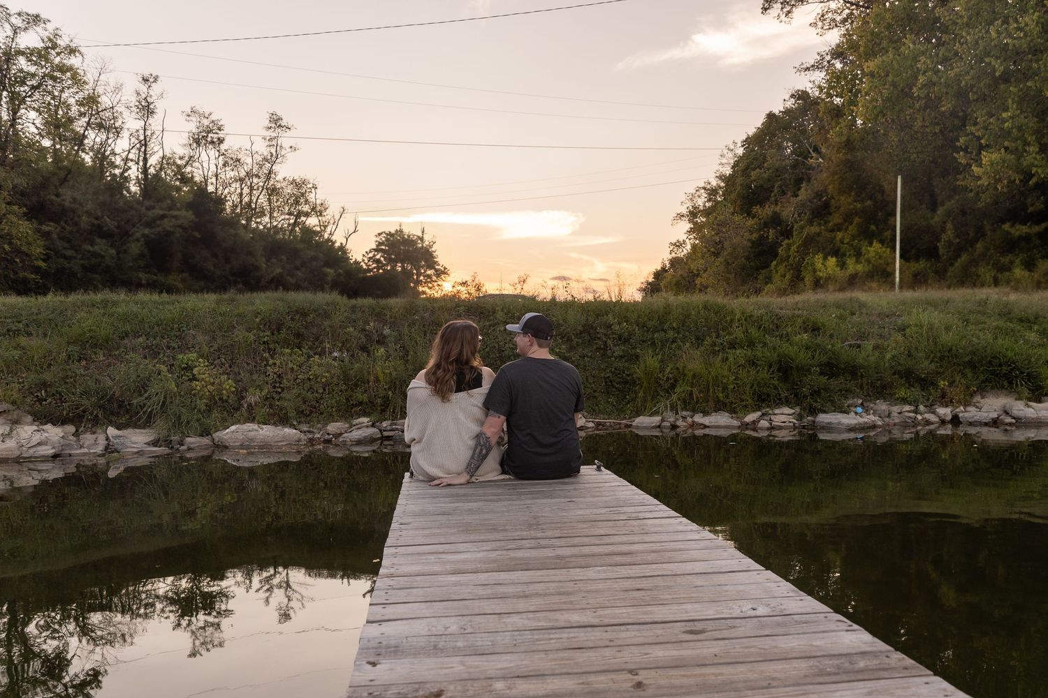 Two people sit on a wooden boardwalk overlooking a serene lake at sunset while surrounded by trees.