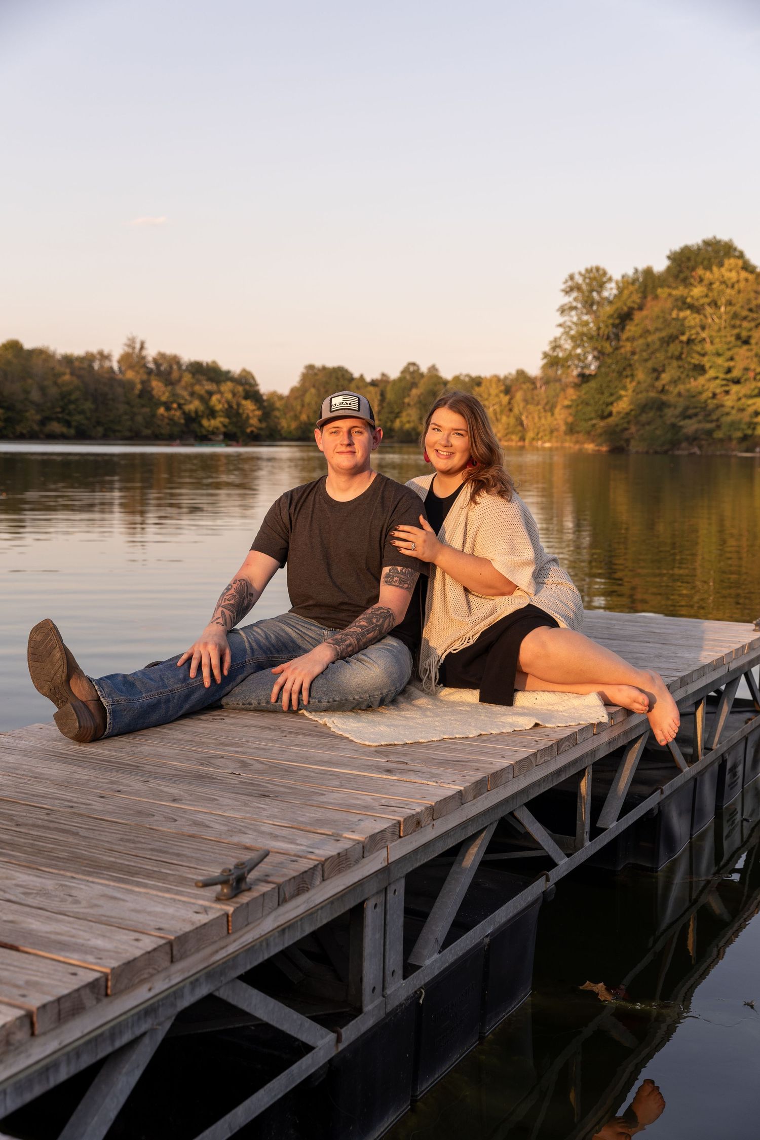 A couple relaxes together on a wooden dock by a serene lake during sunset.