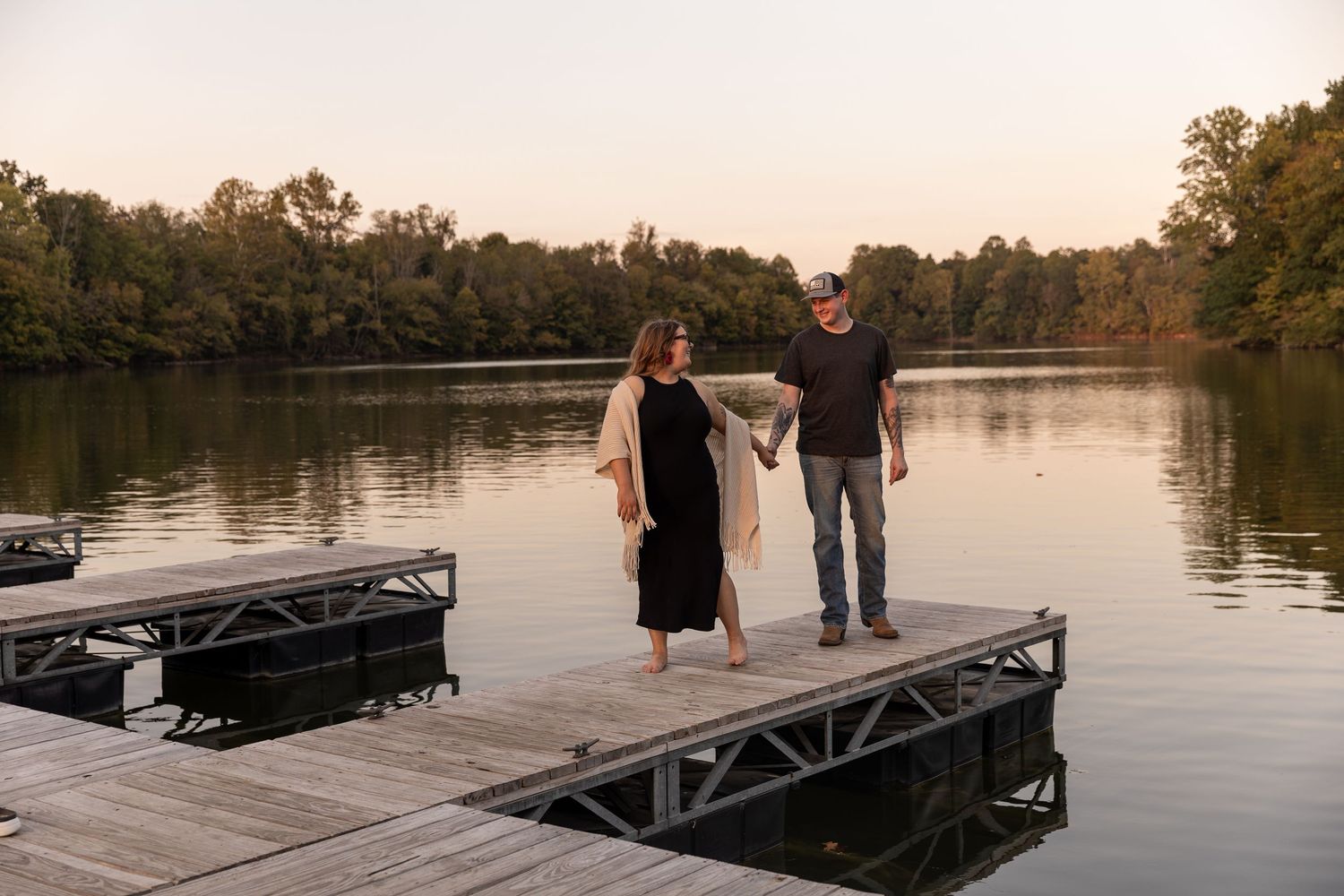 People walking on a wooden dock over a serene lake at sunset with trees in the background.