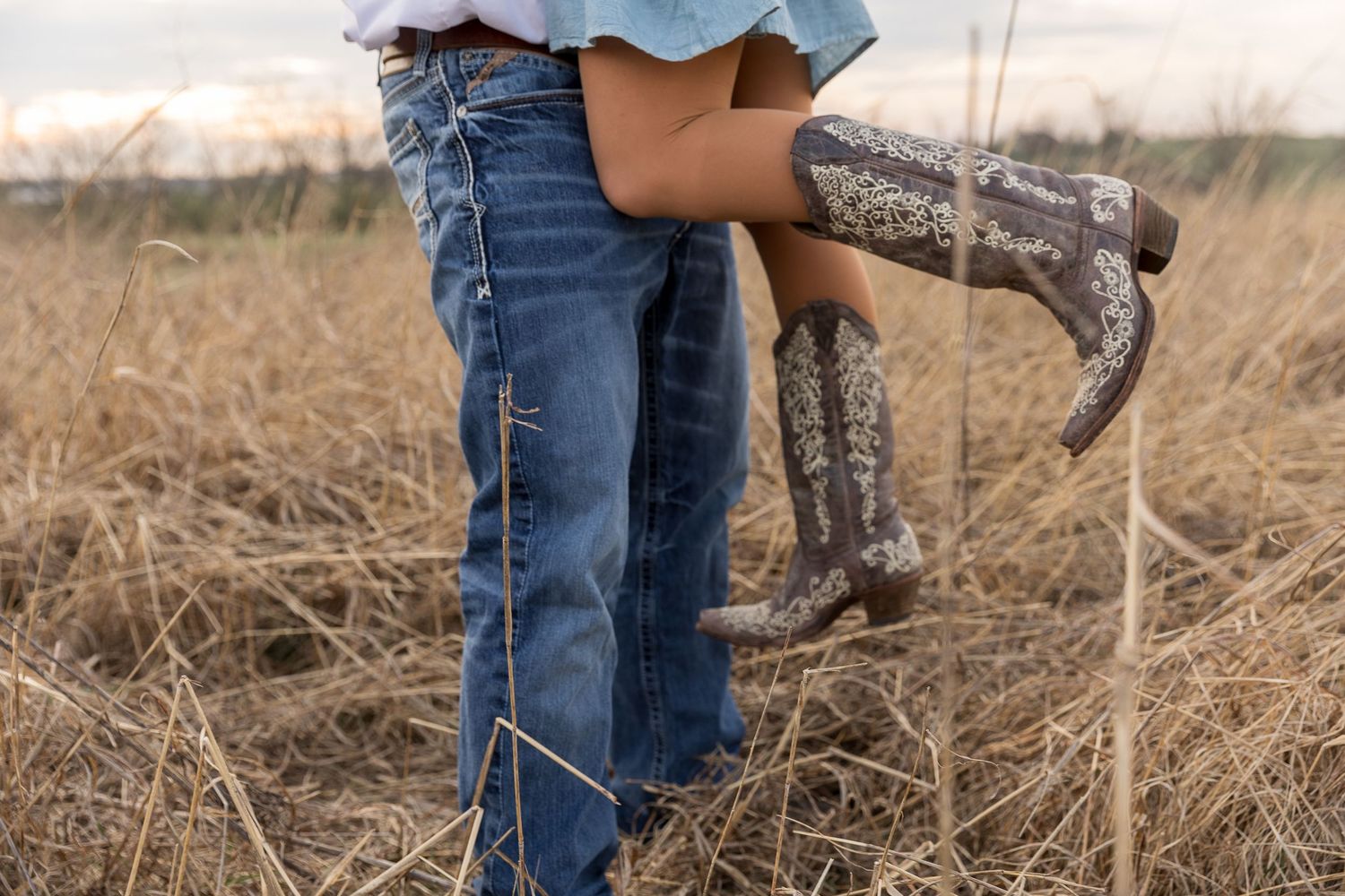 A person wearing brown cowboy boots walking in a field of dried grass and hay on a cloudy day.