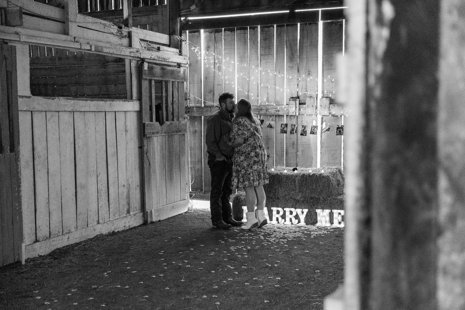 A couple stands together in a dimly lit area with illuminated 'Marry Me' text in the background.