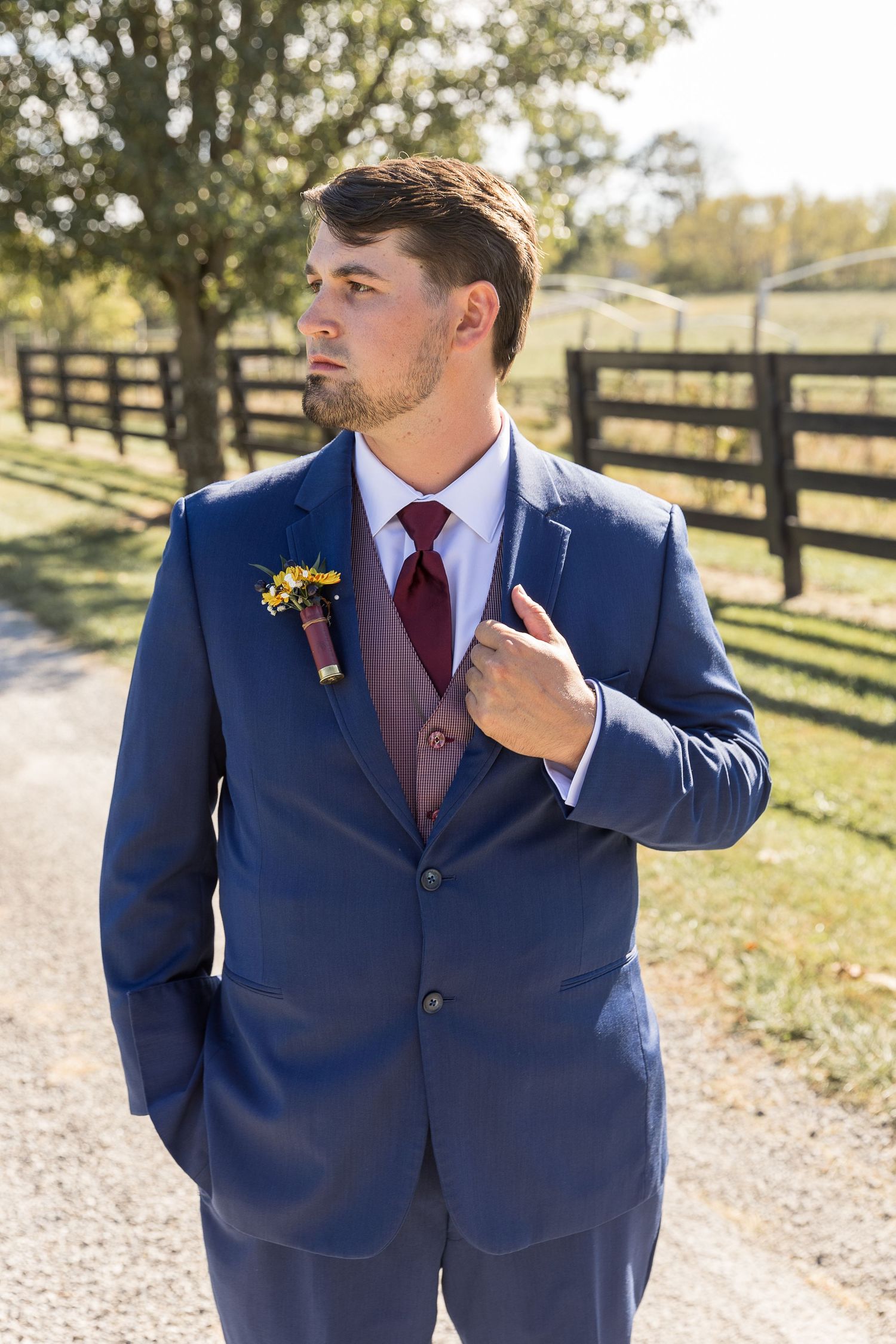 Formal navy suit with burgundy tie and rustic boutonniere against farm fence backdrop.