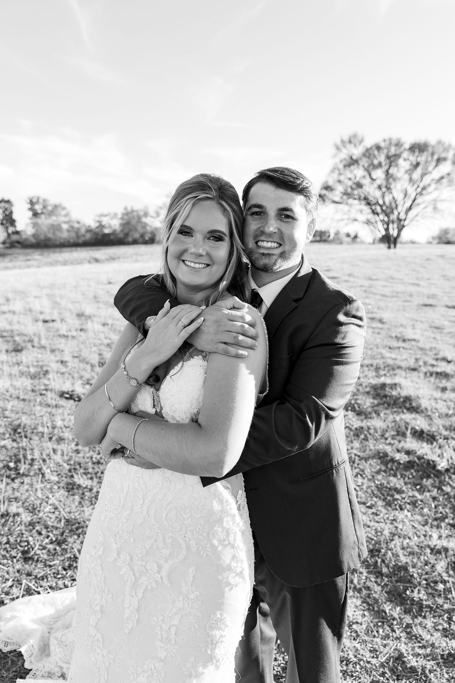A newlywed couple embraces in a sunny field wearing wedding attire in black and white.
