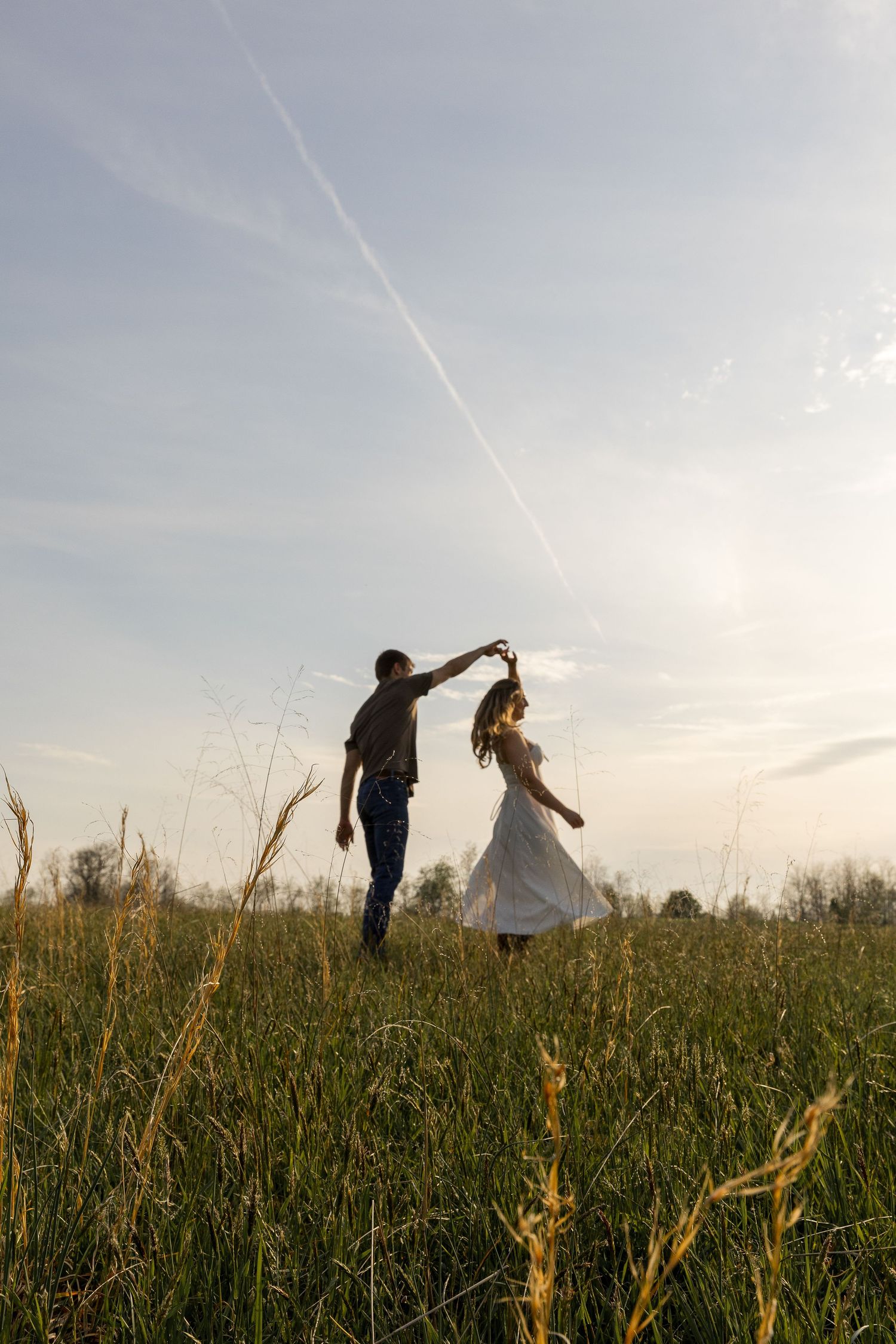 Silhouettes of a couple dance together in a grassy field at sunset with contrails in the sky.