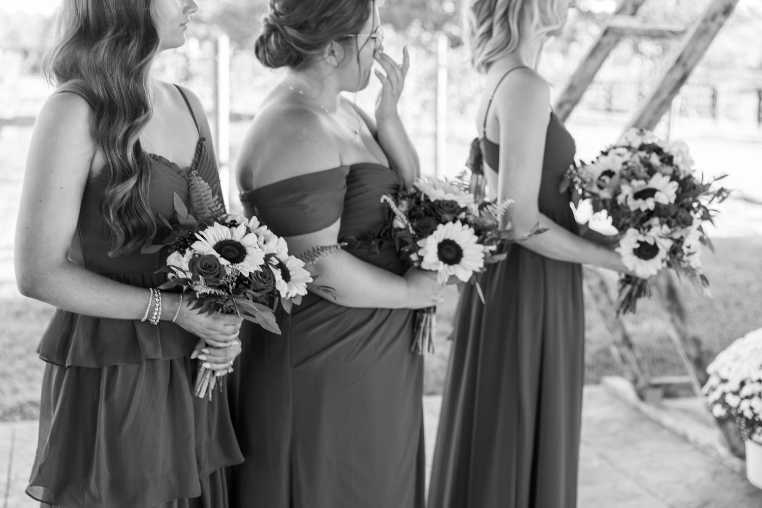 Bridesmaids in dark dresses hold sunflower bouquets at outdoor wedding ceremony.