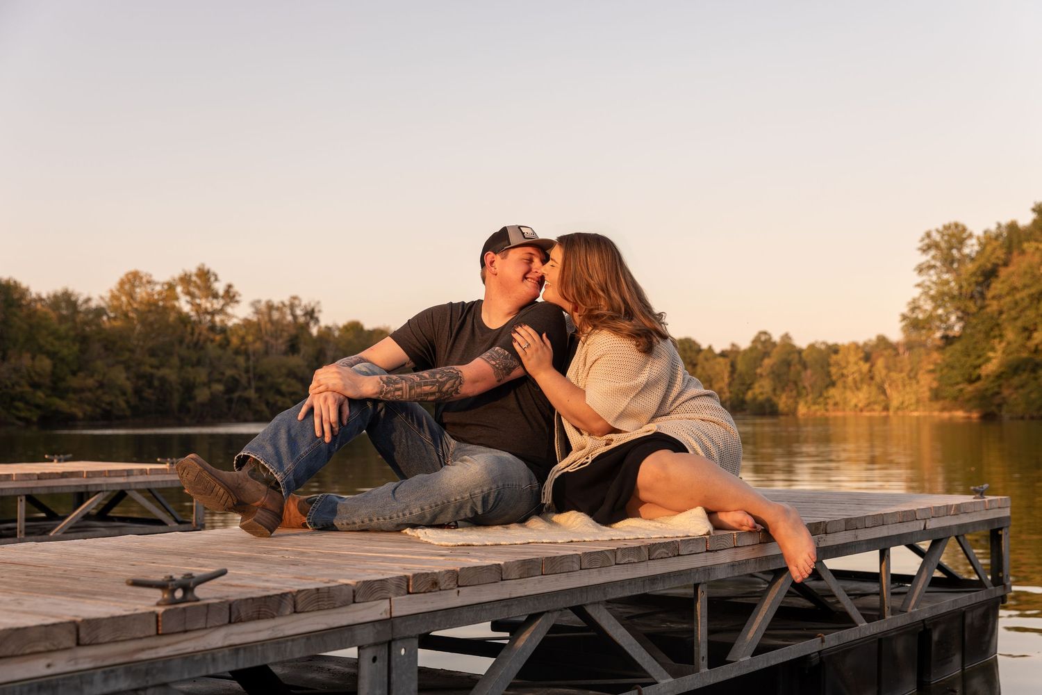 Romantic couple lying and embracing on rustic dock overlooking peaceful lake at sunset.